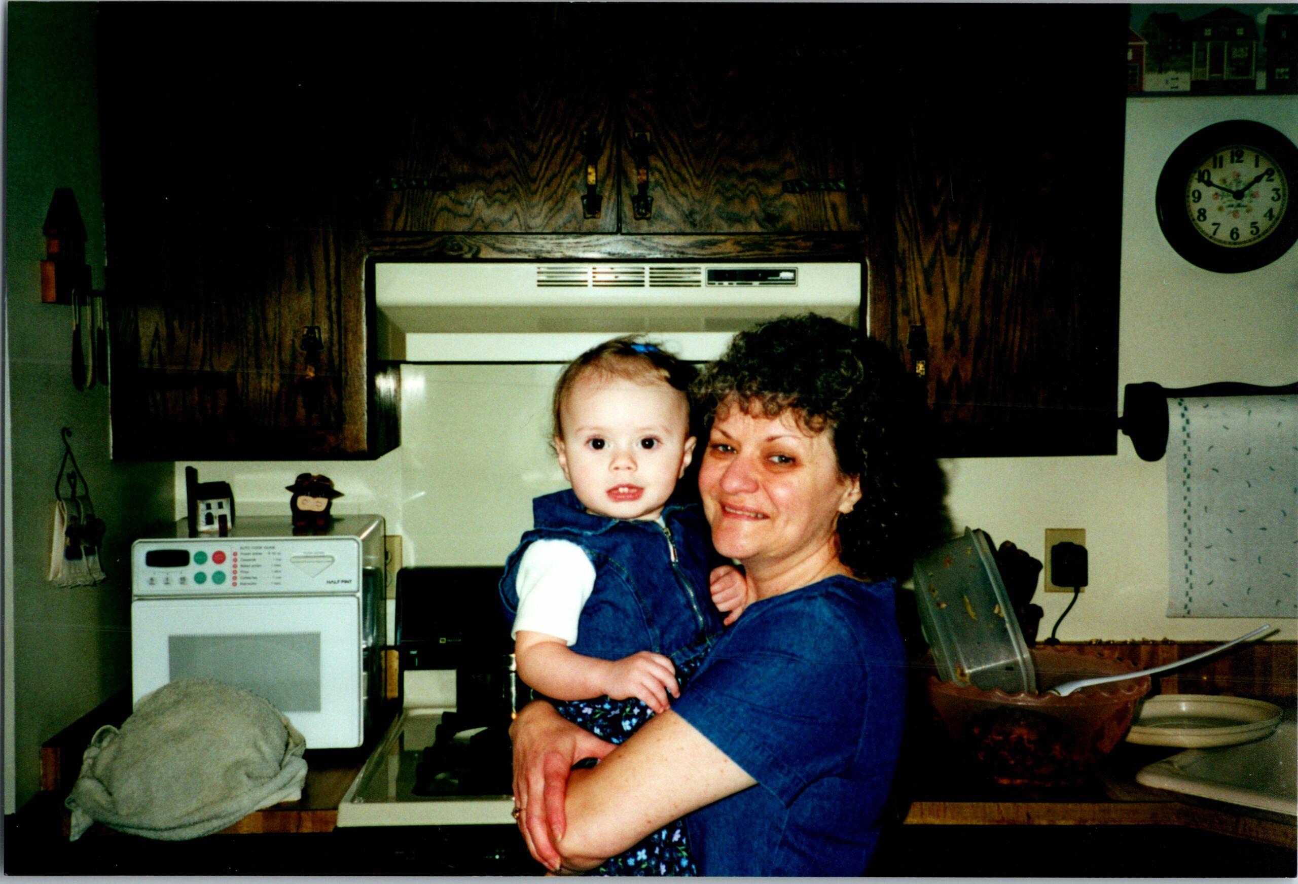 Woman holding a baby in a kitchen, with cabinets, stove, and a wall clock in the background.