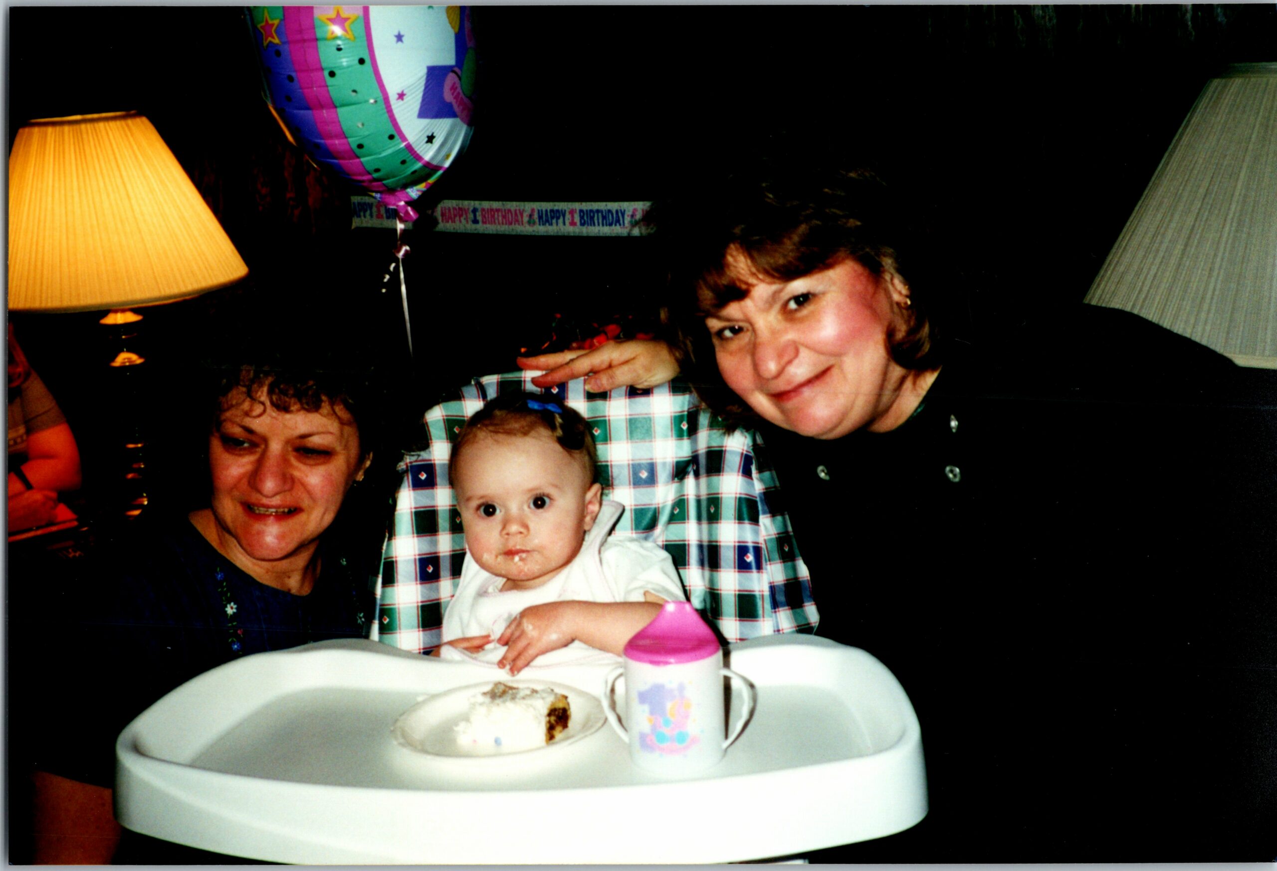 Baby in a high chair at a birthday party, flanked by two smiling adults; cake and a pink-and-white cup on the table with balloons in the background.