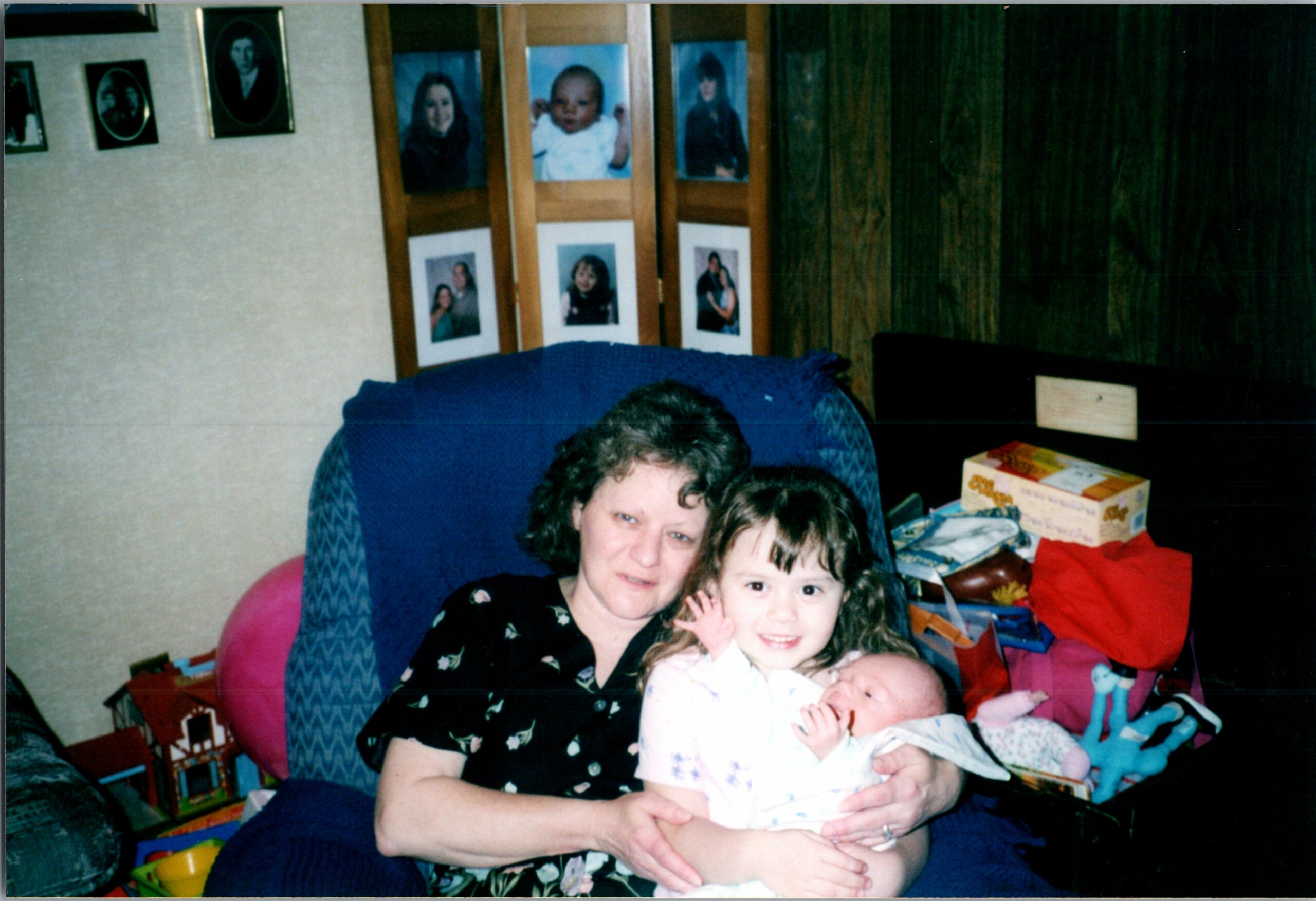 Woman with two babies on her lap in a living room, surrounded by wall photos and toys in the background.