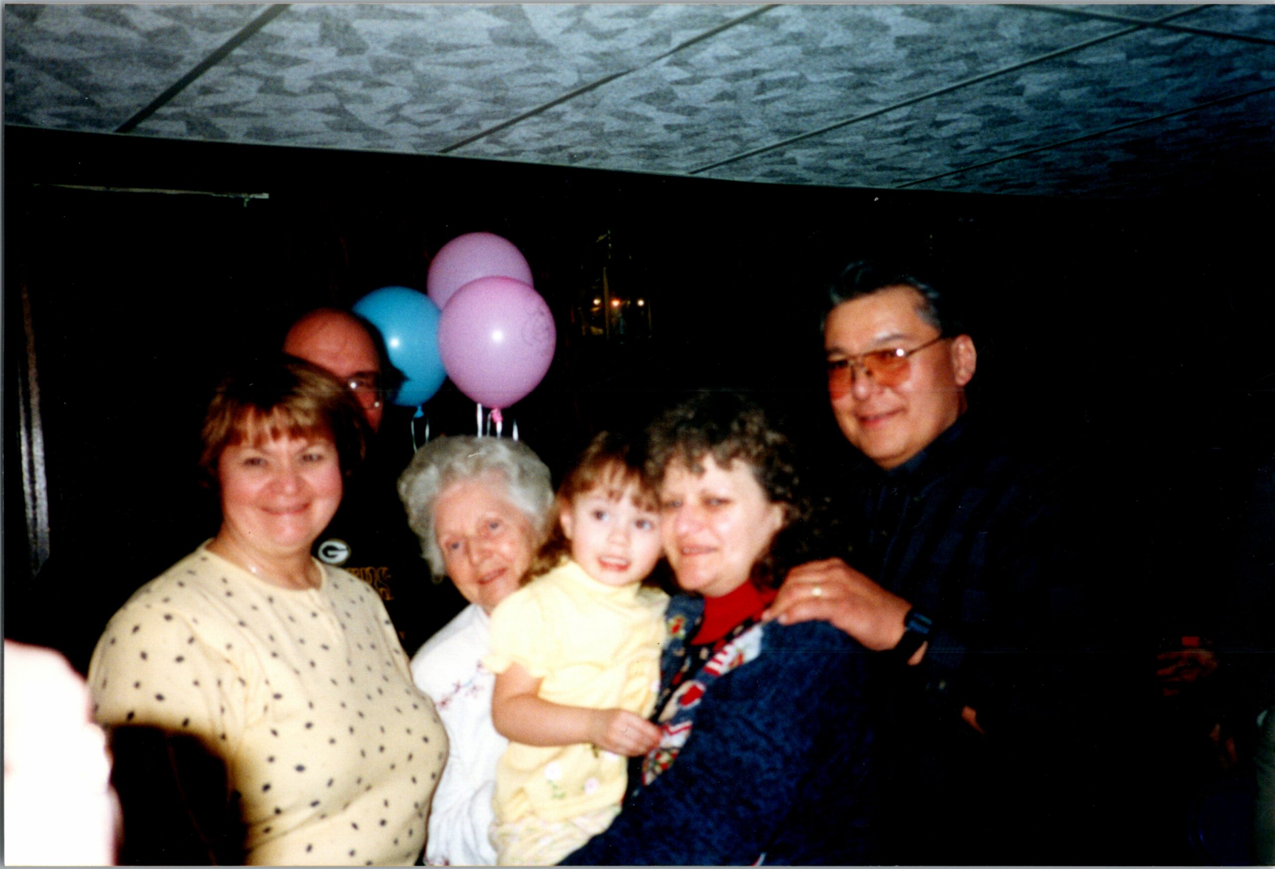 Group of people posing at a celebration, a woman in a light polka-dot sweater on the left, a child in yellow, pink and blue balloons in the background.