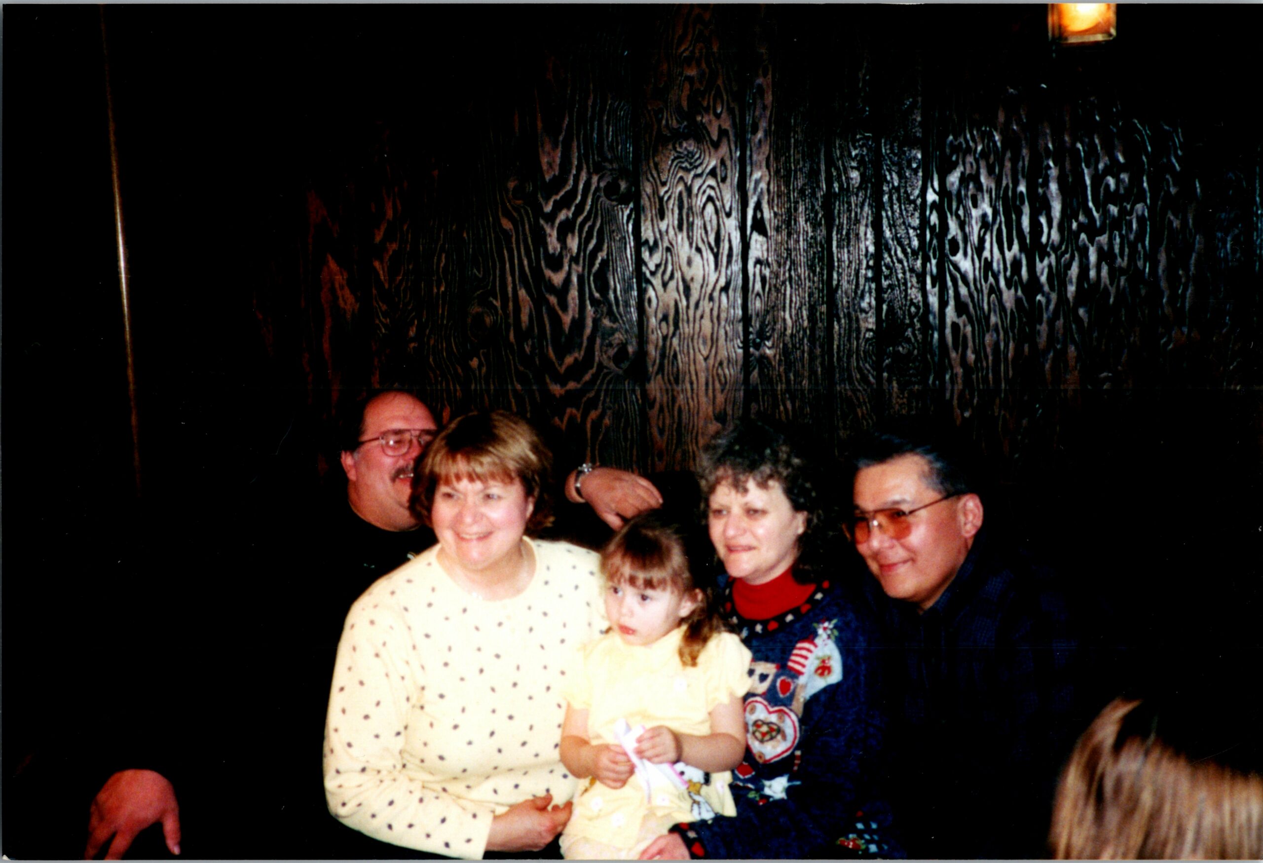 Group of adults and a child posing for a photo at a gathering; woman in a cream polka-dot sweater holds a young child in yellow front center.