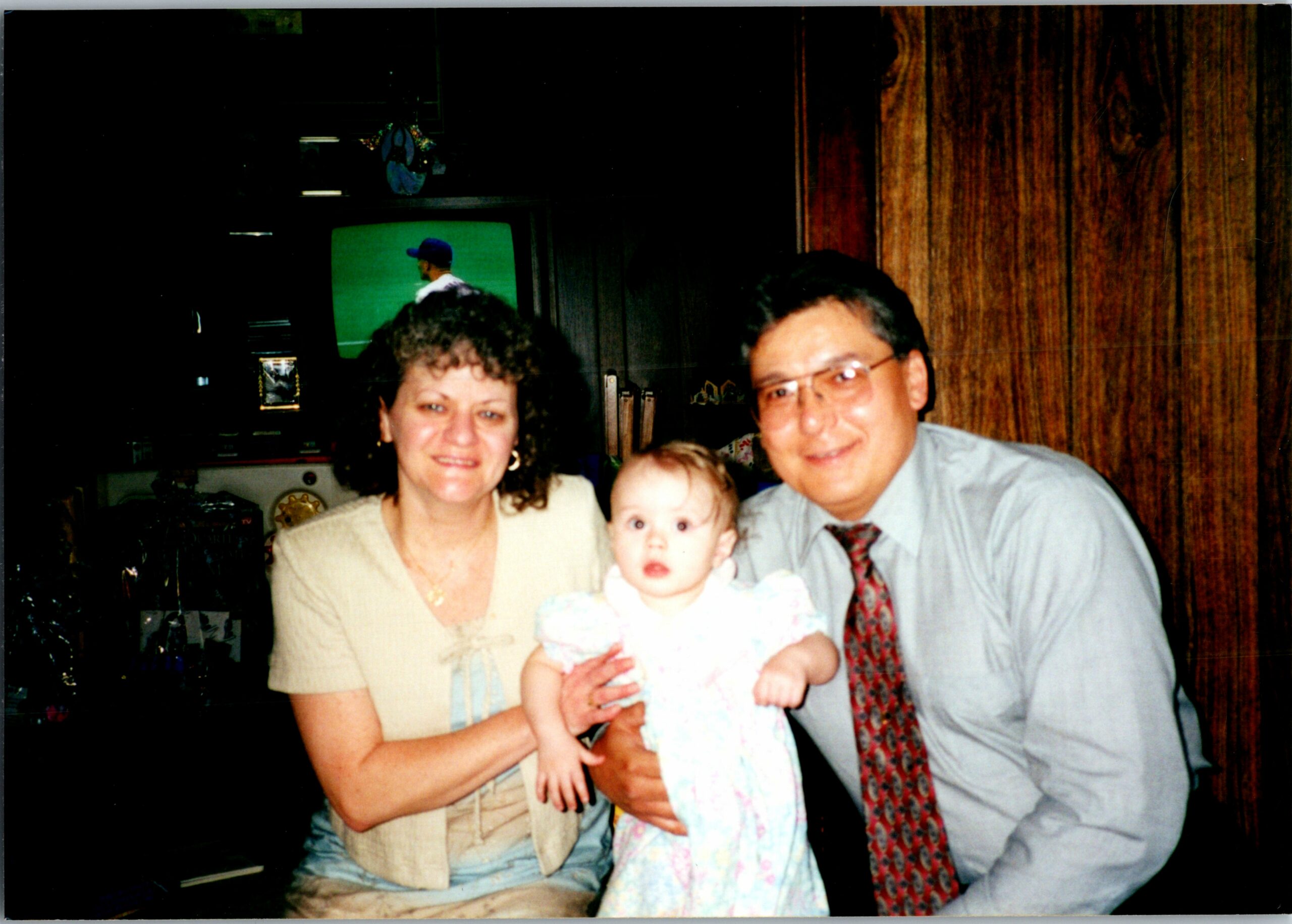 Smiling woman and man holding a baby together indoors, with dark wood paneling in the background.