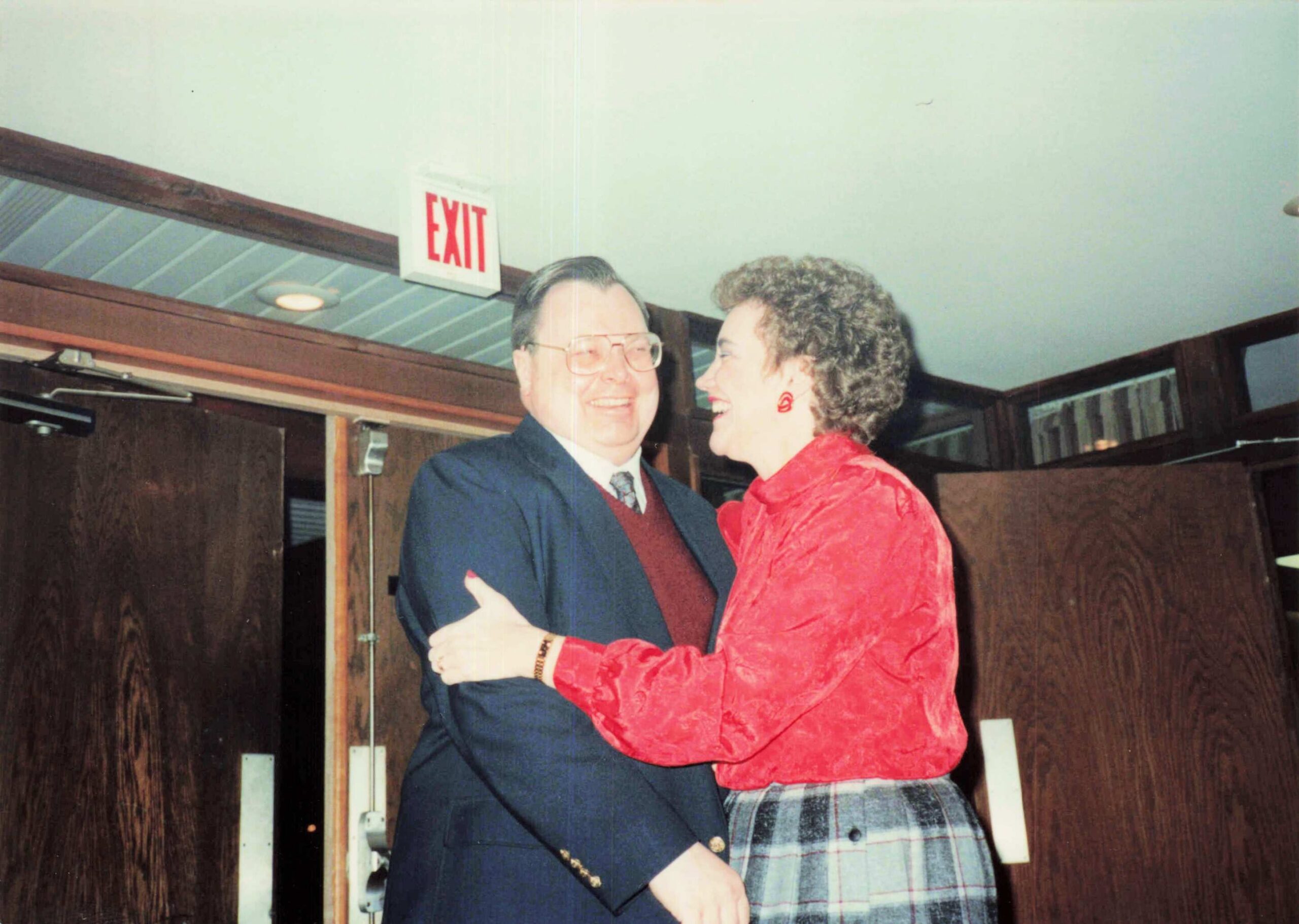 Two people hugging in a doorway inside a building, a man in a suit and a woman in a red jacket smiling at each other under an EXIT sign.