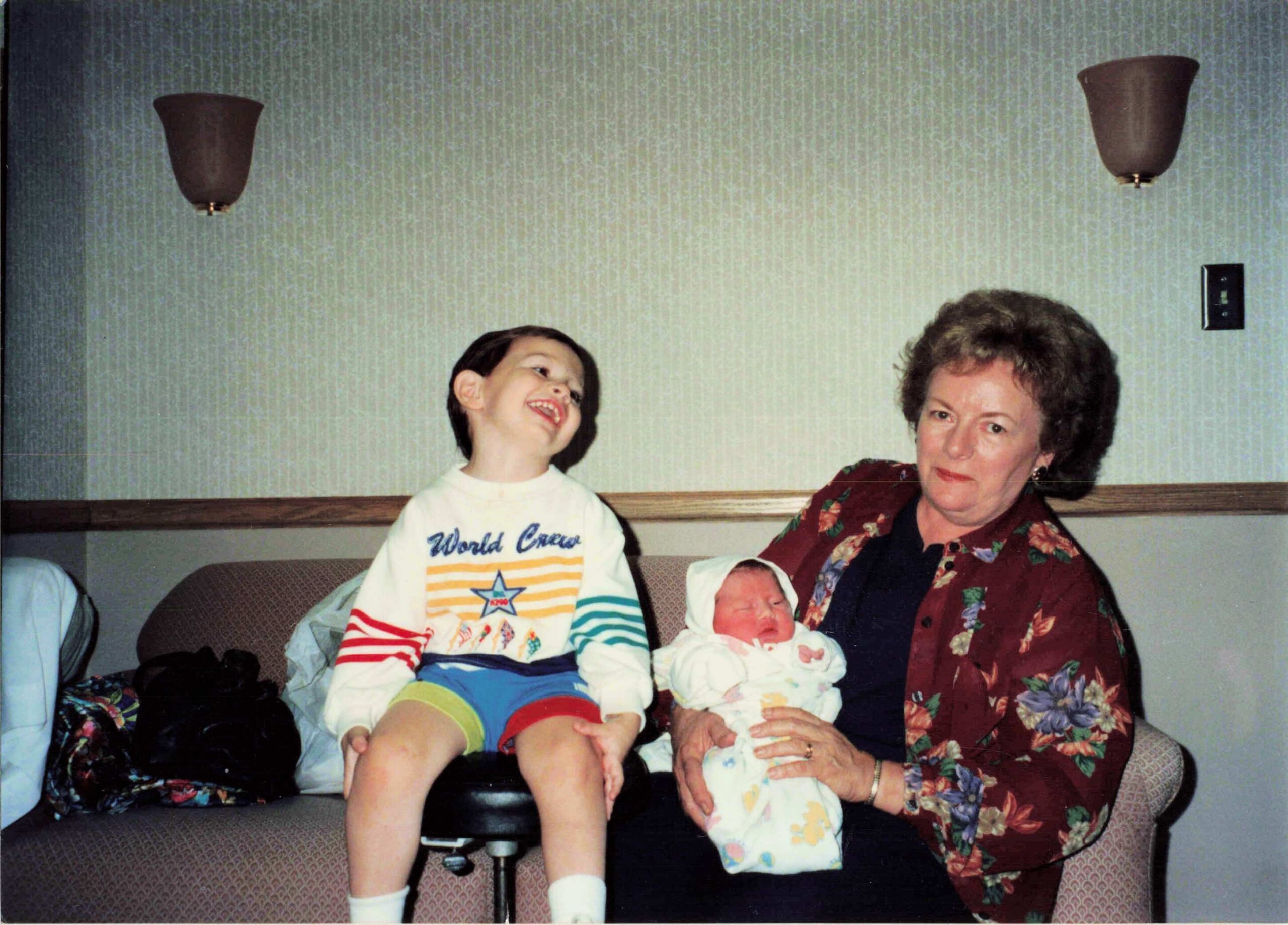 A woman on a sofa with two babies: one baby in her arms and a smiling toddler sitting beside her, in a living room with patterned upholstery.