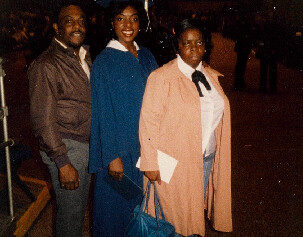 Three adults pose for a photo at a formal event: a man in a brown suit, a woman in a blue gown, and another woman in a peach cape holding a white envelope.