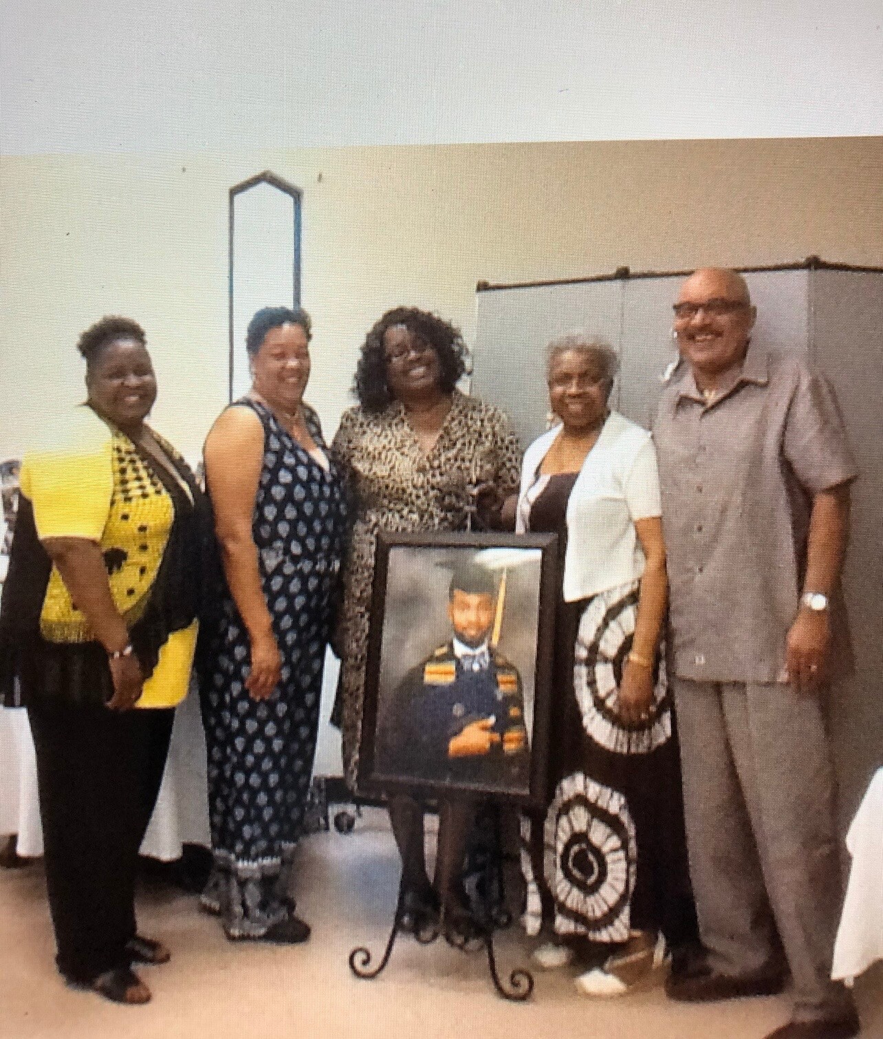 Six adults stand together smiling, with a framed portrait of a man in military uniform displayed on a stand in front of them.