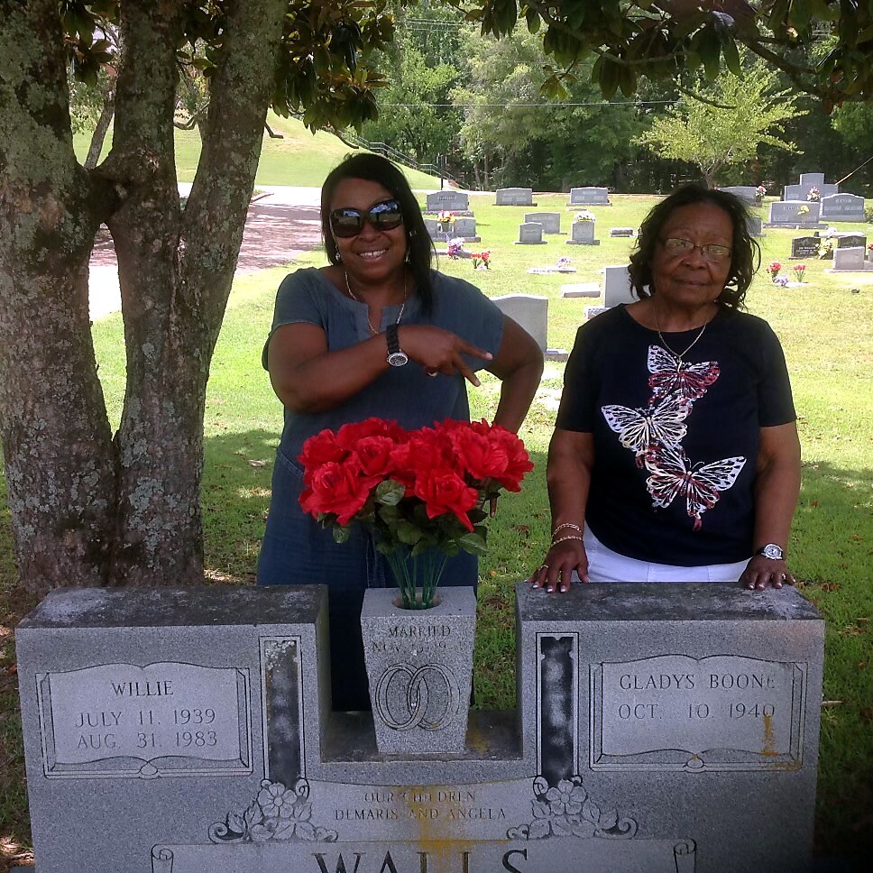 Two women stand behind a family grave marker under a tree; one points to the vase of red roses between the two tombs, with headstones in a cemetery backdrop.