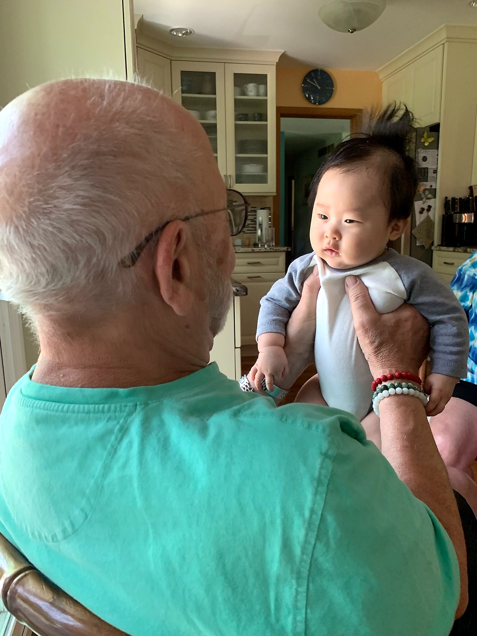 Older man with gray hair and glasses facing a baby being held up in a bright kitchen.