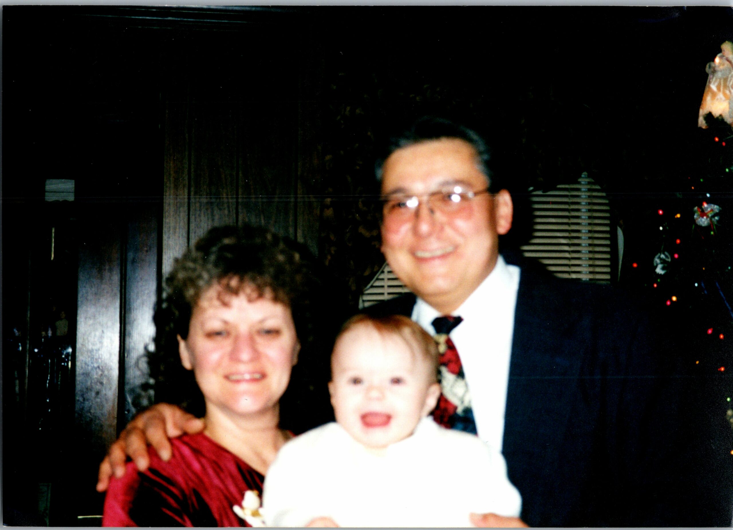 Family group photo at a celebration: woman in red, man in a suit, and a child in the foreground smiling at the camera.