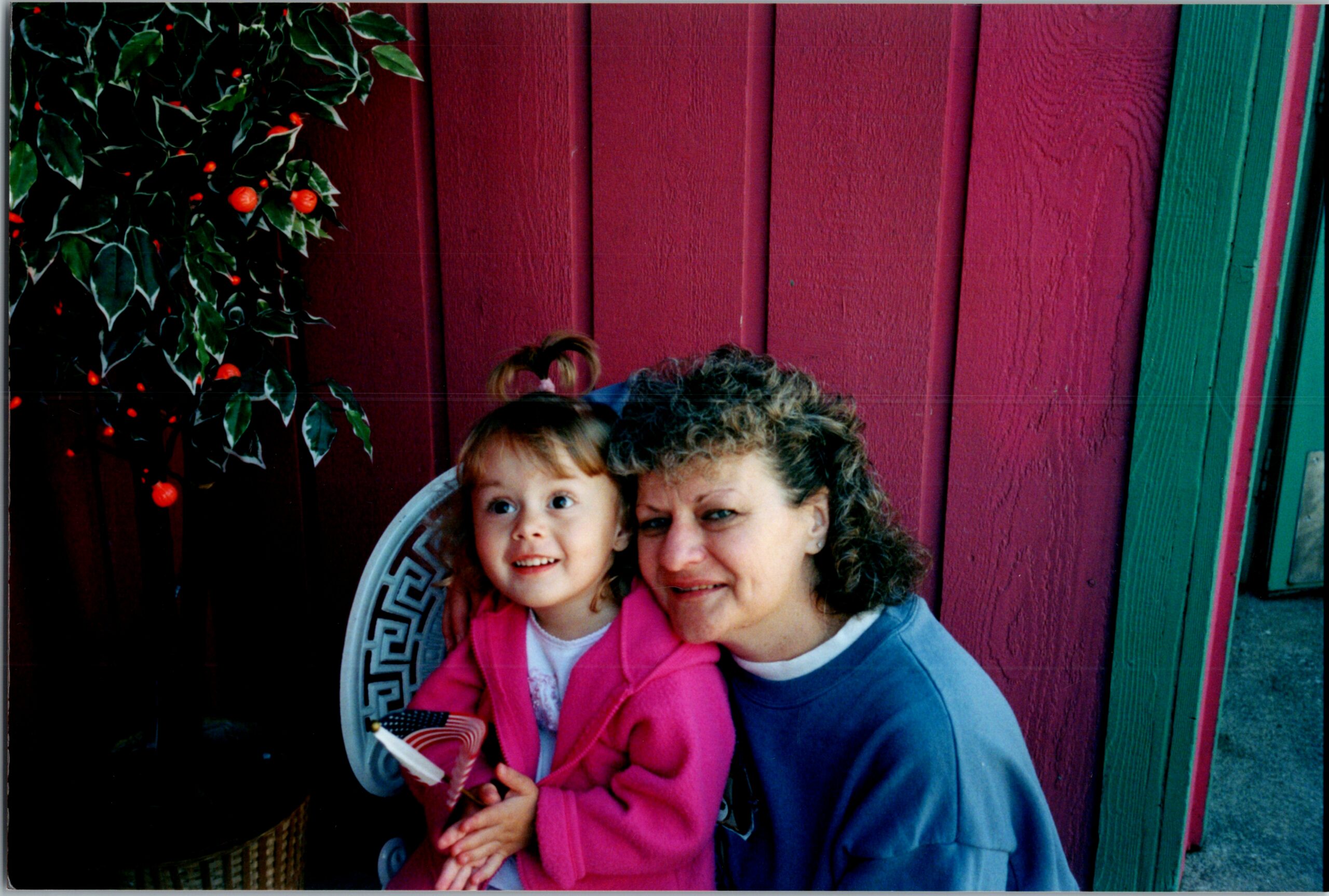 Woman and child sit together outdoors, smiling at the camera in front of a magenta wooden fence with holly and red berries on the left.