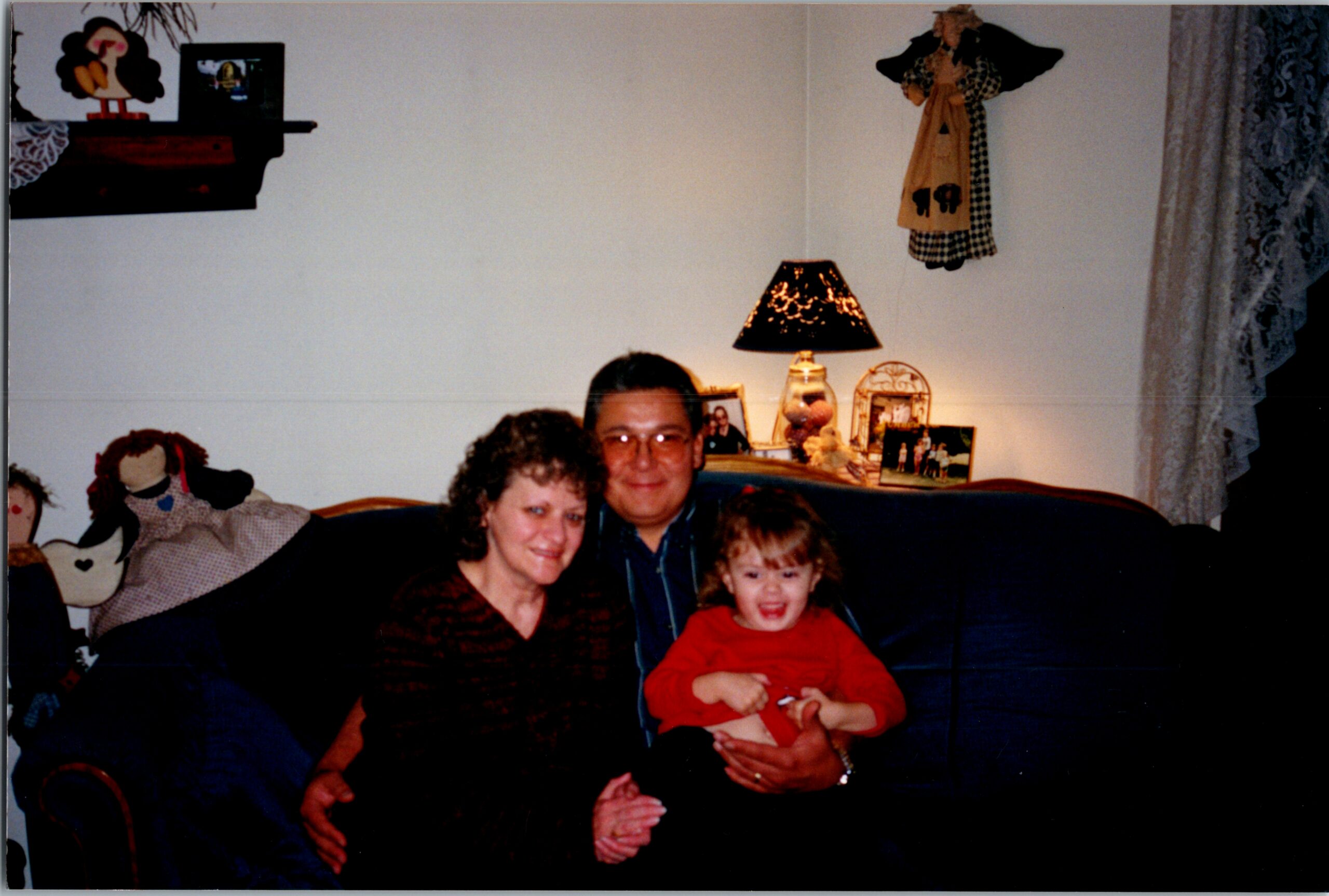 Family sits on a sofa with a smiling toddler in red on the lap of a man and woman in a cozy living room, plus a lit lamp and family photos in the background.