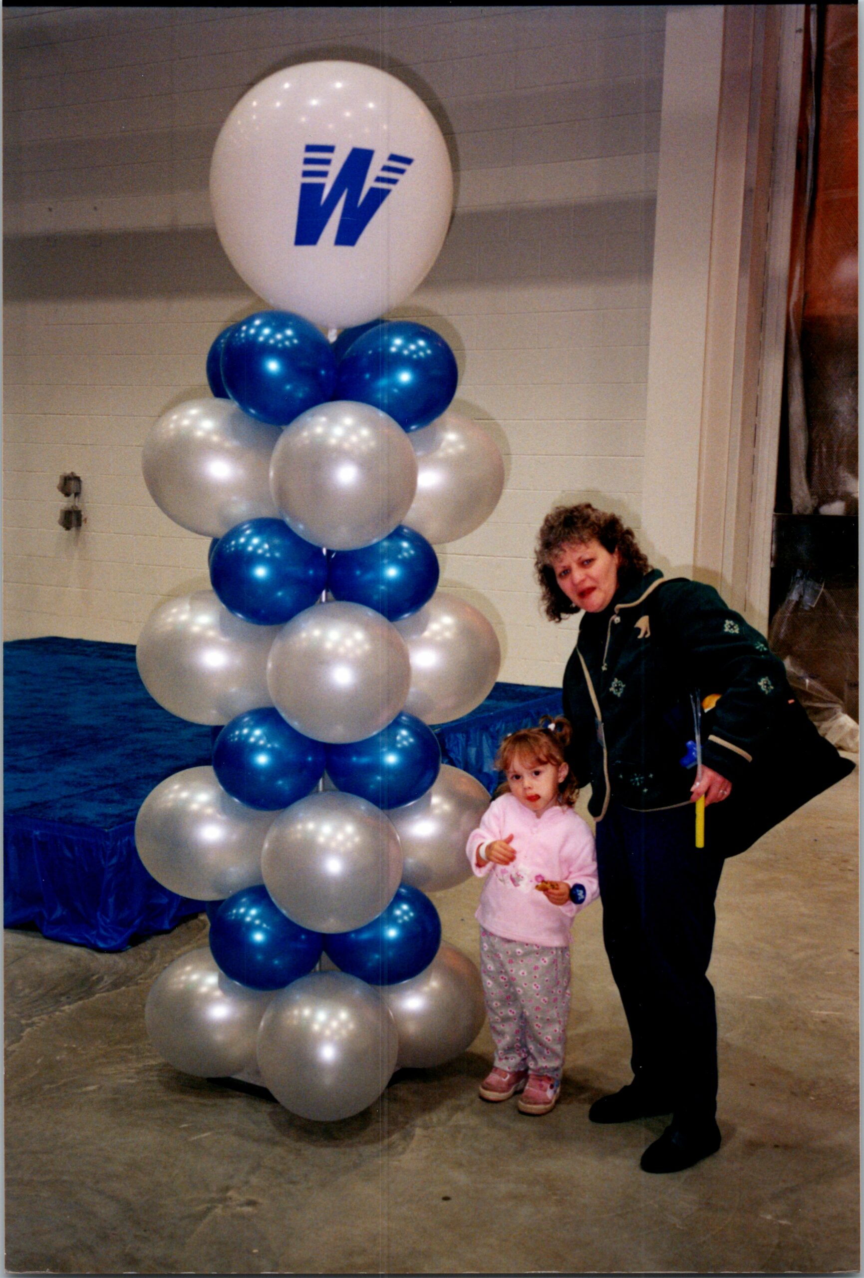 Tall column of blue and silver balloons with a large white top balloon featuring a blue 'W' logo, indoors, with a woman and child nearby.