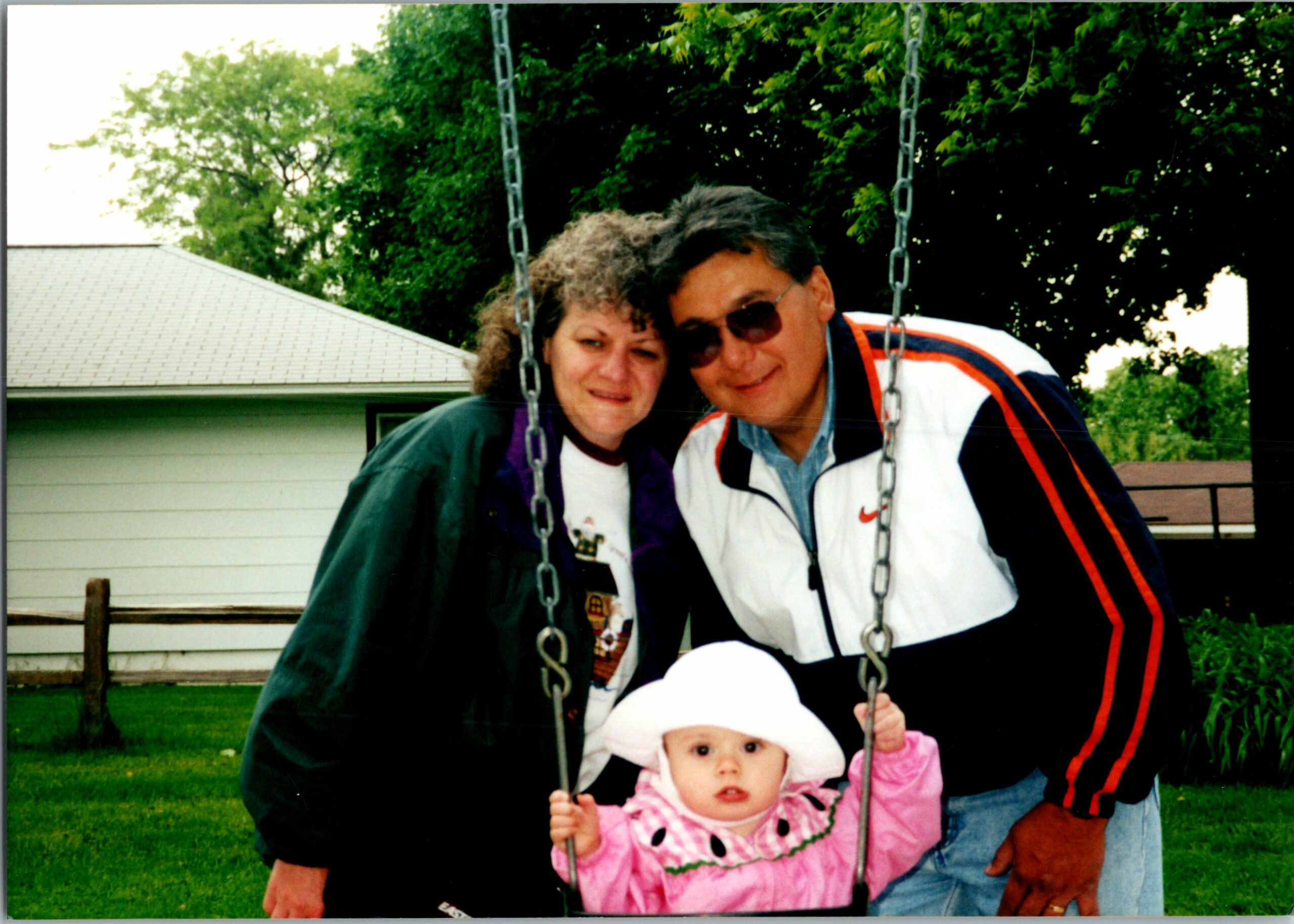 A couple stands behind a baby on a swing in a backyard, with green grass and trees in the background.