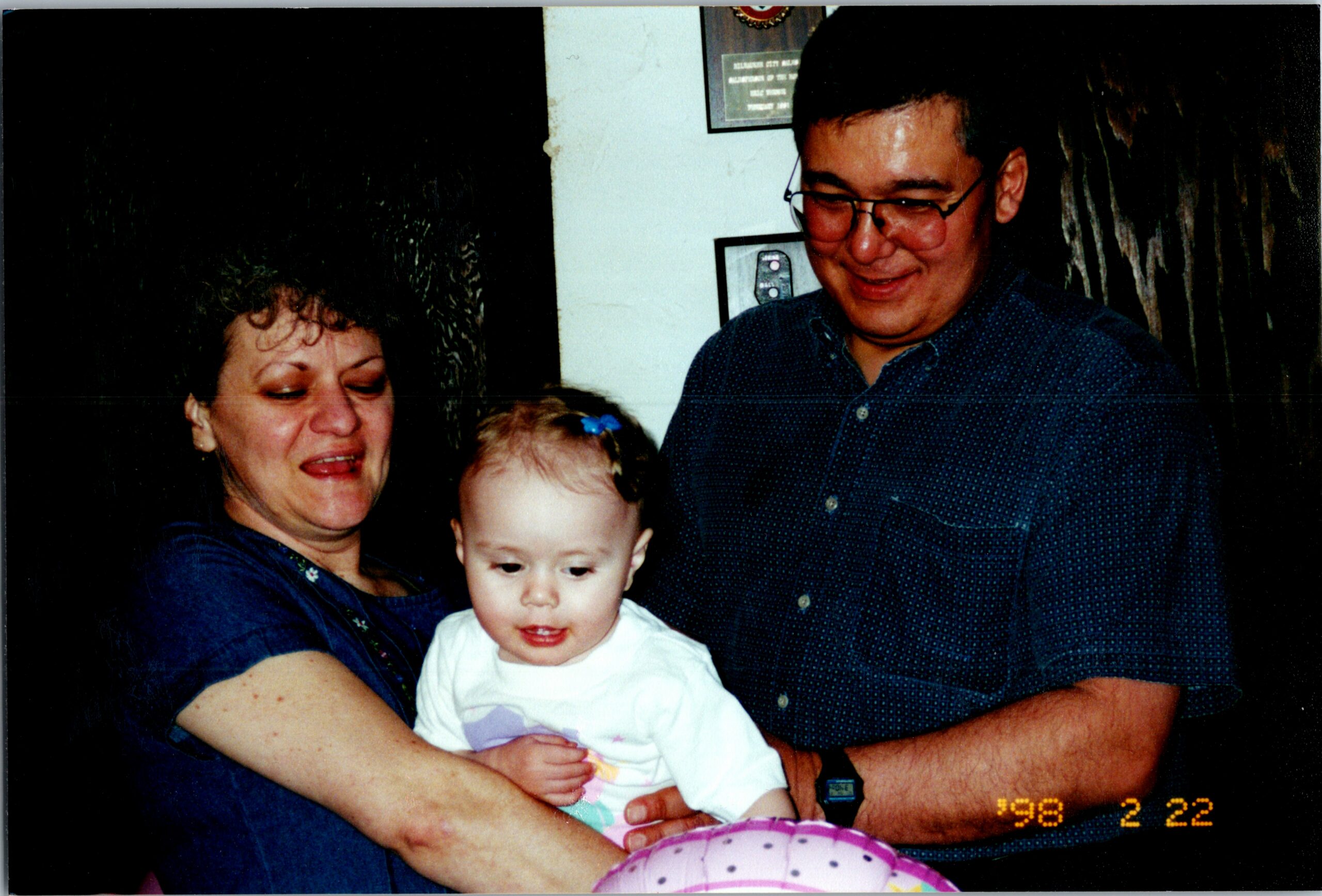 Woman holding a baby, smiling, with a man in glasses beside them in a dimly lit room.