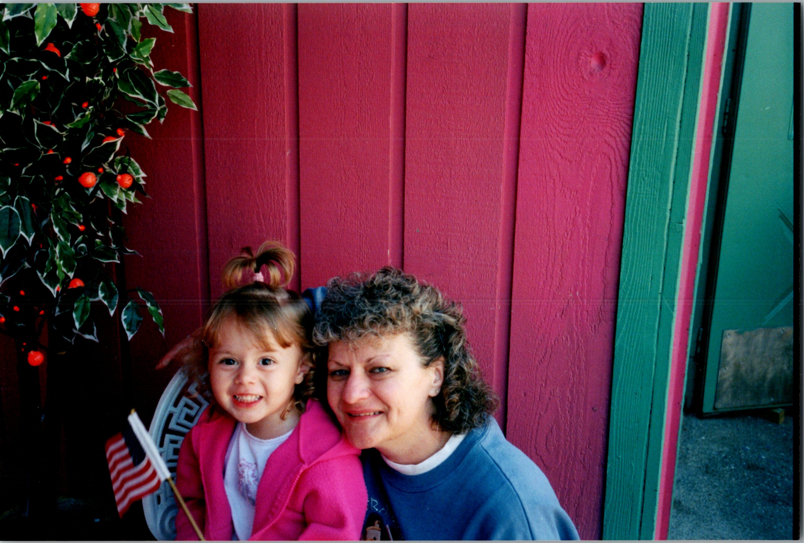 A young girl in a pink jacket and an older woman smile together in front of a bright pink and green wooden wall with festive holly on the left.