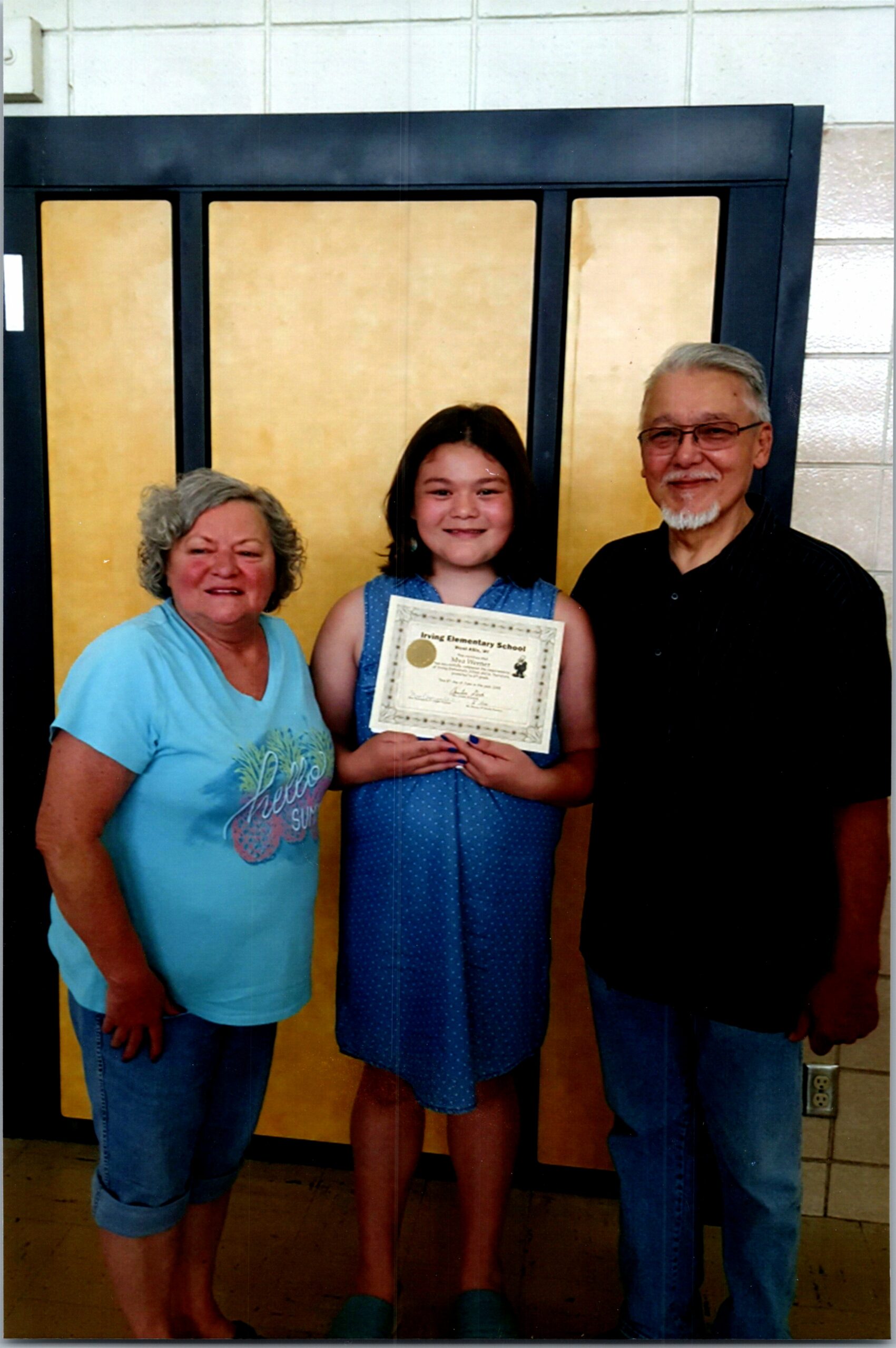 Three people posing indoors: an elderly woman in a light blue shirt, a smiling girl in a blue dress holding a certificate, and a man with glasses next to her.