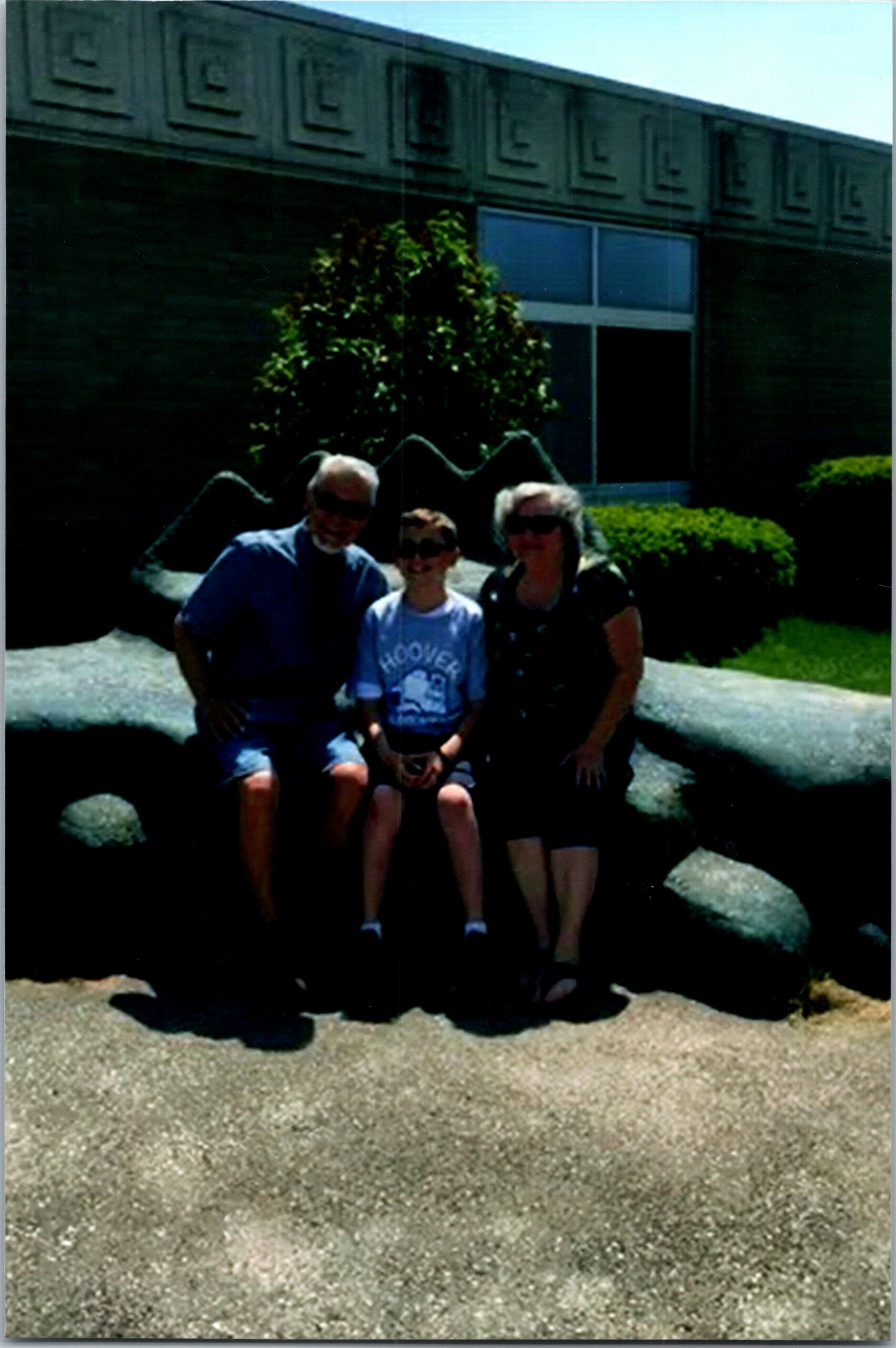 Family of three posing on a rock bench in front of a dark brick building with a window in daylight.