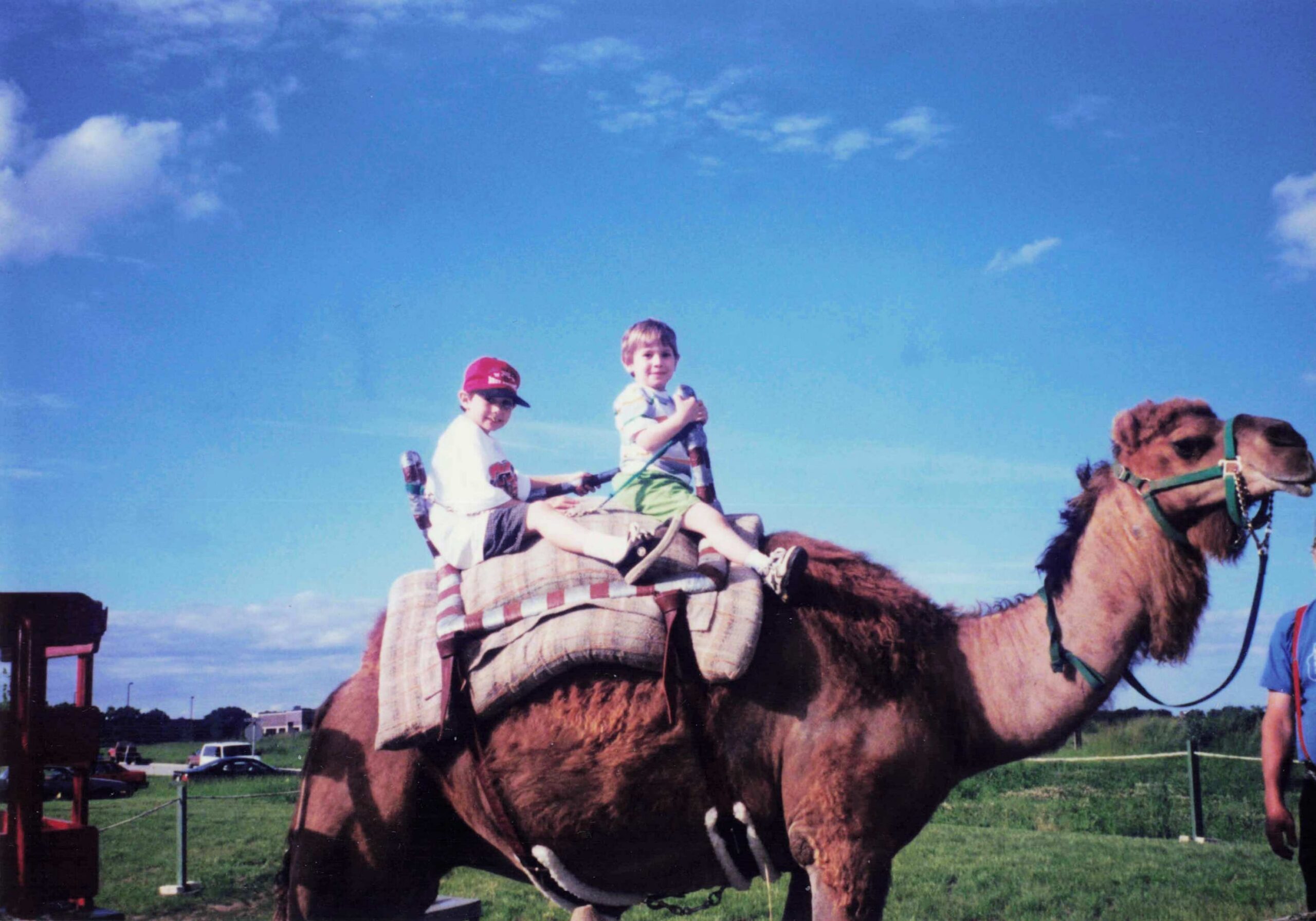 Two children ride a camel together on a sunny day, sitting on a padded saddle blanket with a blue sky above.
