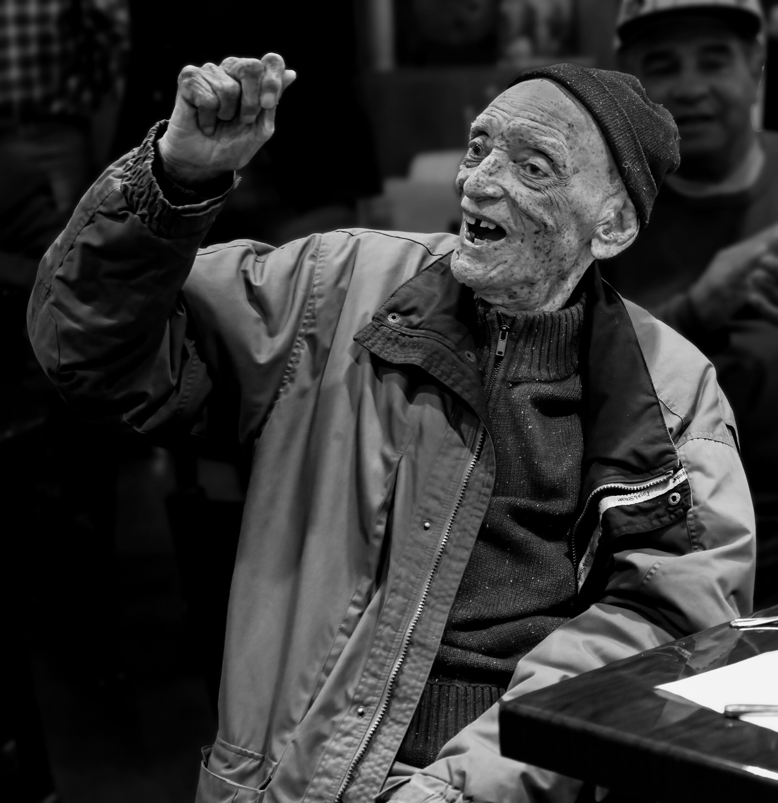 Black-and-white portrait of an older man in a beanie and jacket, raising a clenched fist in a celebratory gesture.
