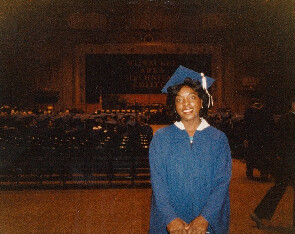 Graduate in a blue cap and gown posing for a photo at a graduation ceremony with a dark auditorium backdrop behind her.