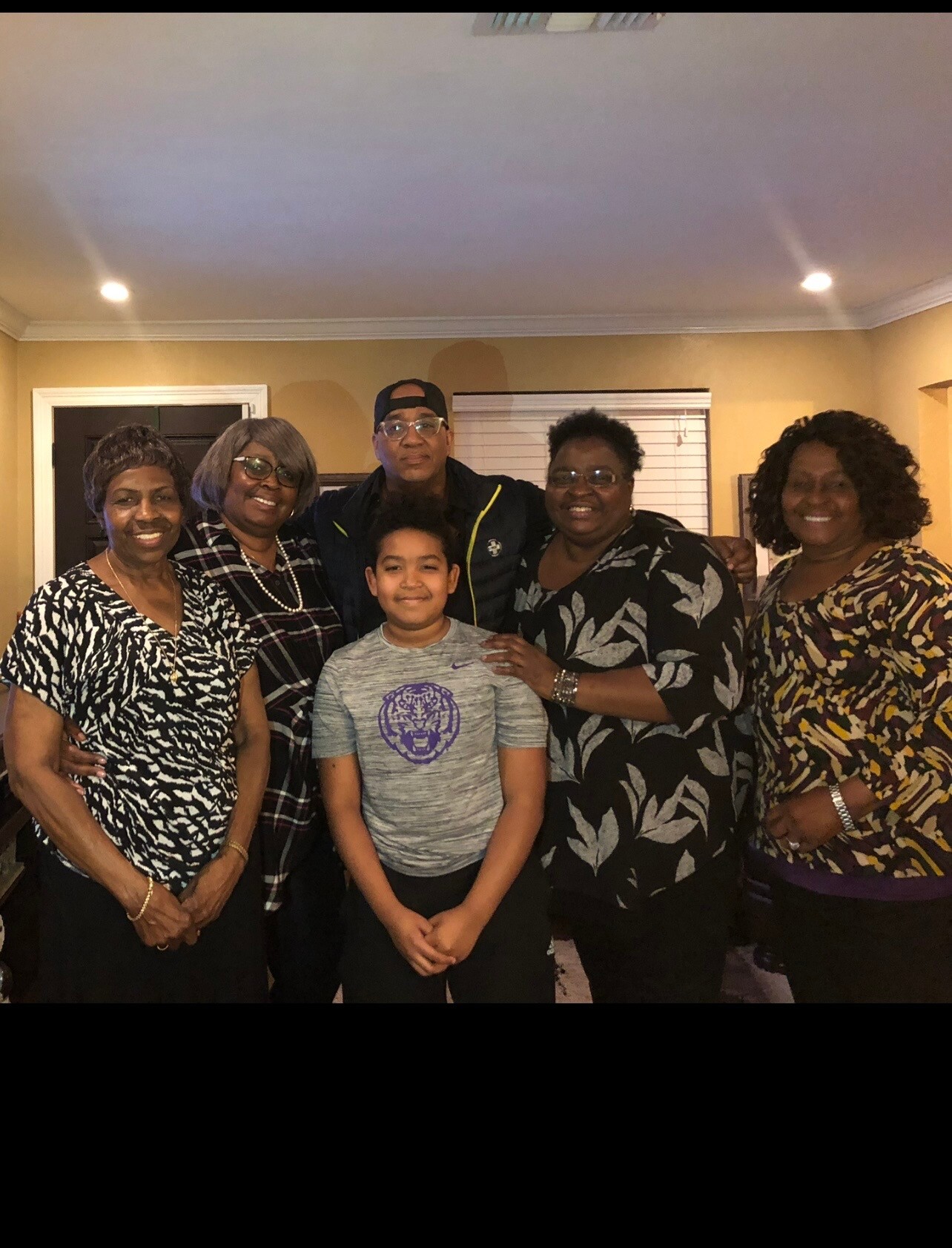 A family group posing in a living room, six adults and one child smiling at the camera with warm wall tones in the background.