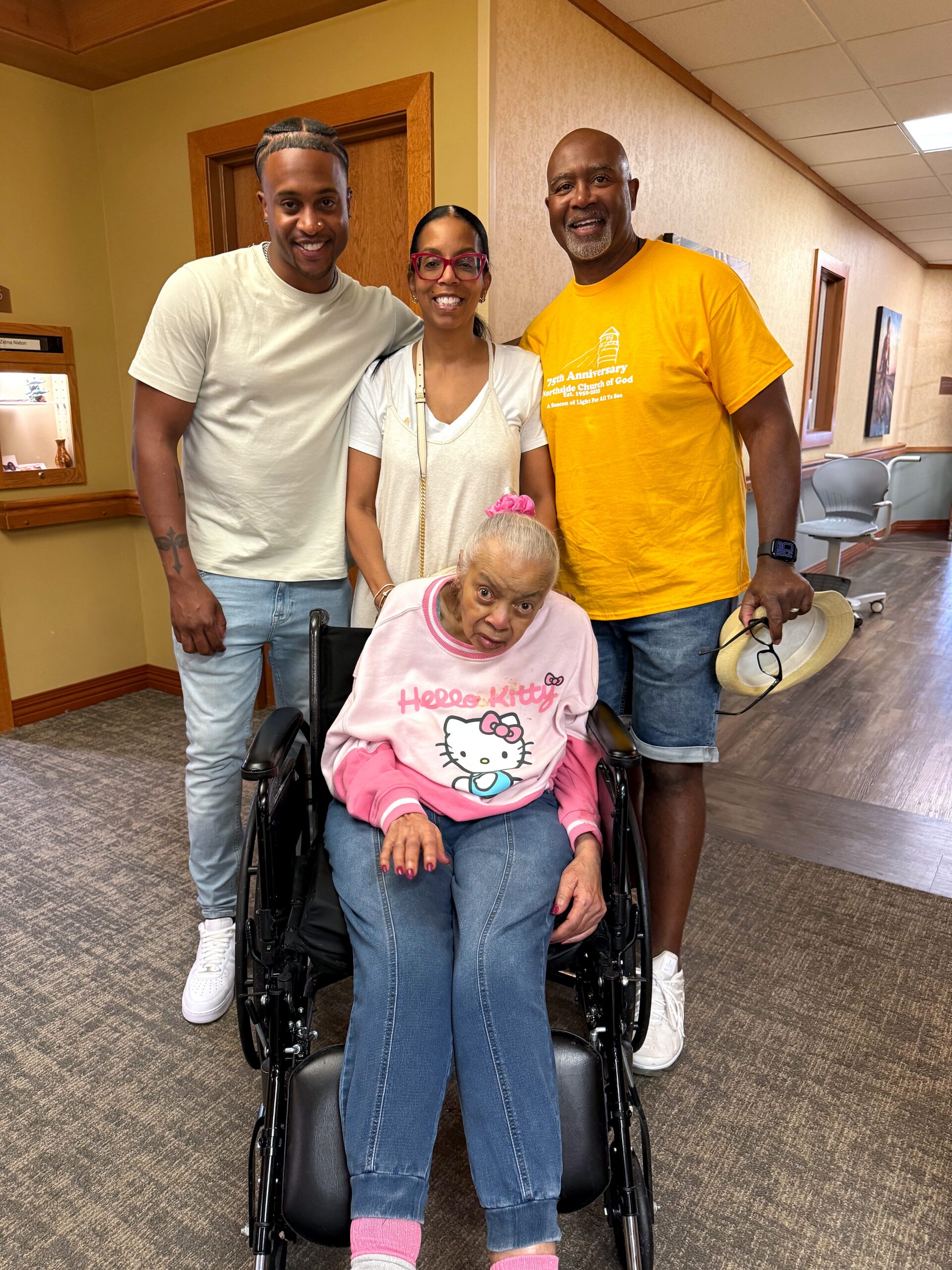 Group of four people posing in a hallway, with a smiling elderly woman in a wheelchair in front.