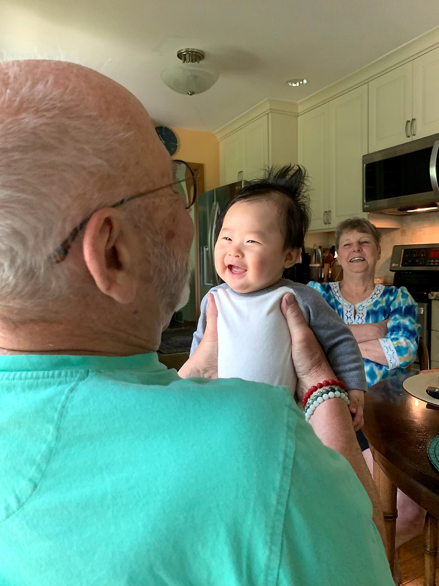 Baby being held by adults in a bright turquoise shirt foreground; smiling woman in a blue patterned blouse in a kitchen.