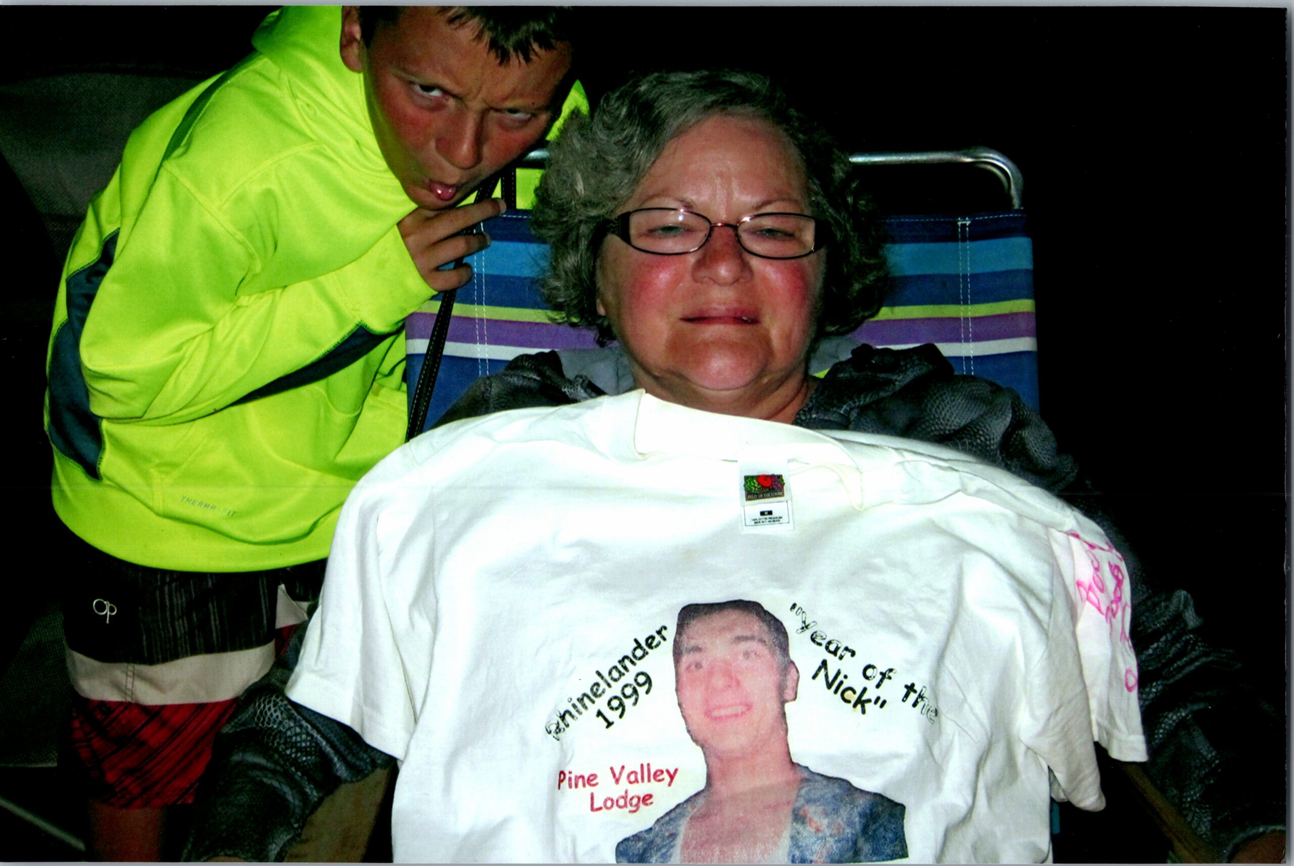 Boy in a neon yellow jacket leans close to an older woman with glasses seated in a striped outdoor chair at night.
