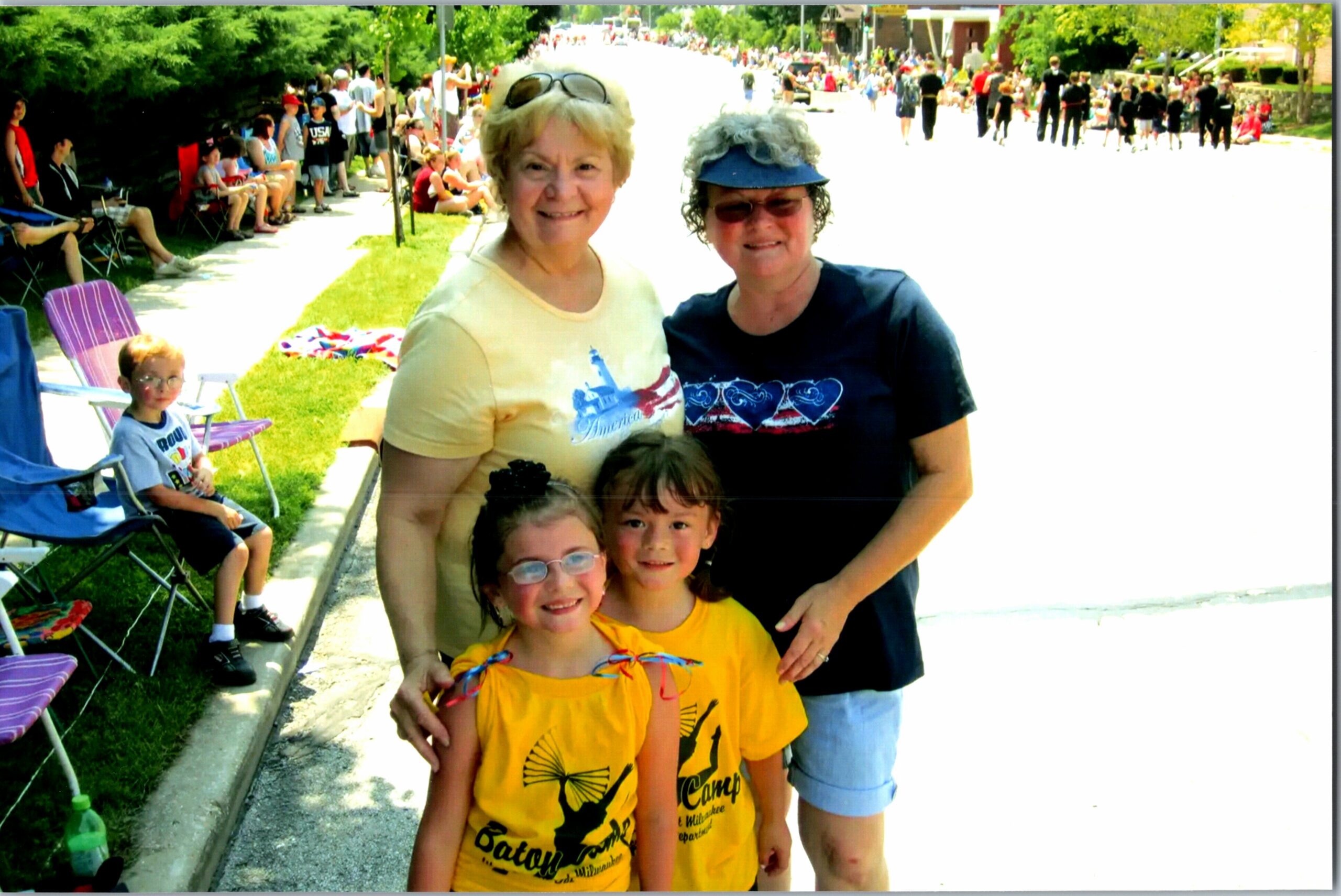 Smiling group of five (three kids in yellow shirts and two adults) posing at a sunny daytime street parade, with spectators seated along the curb on the left.