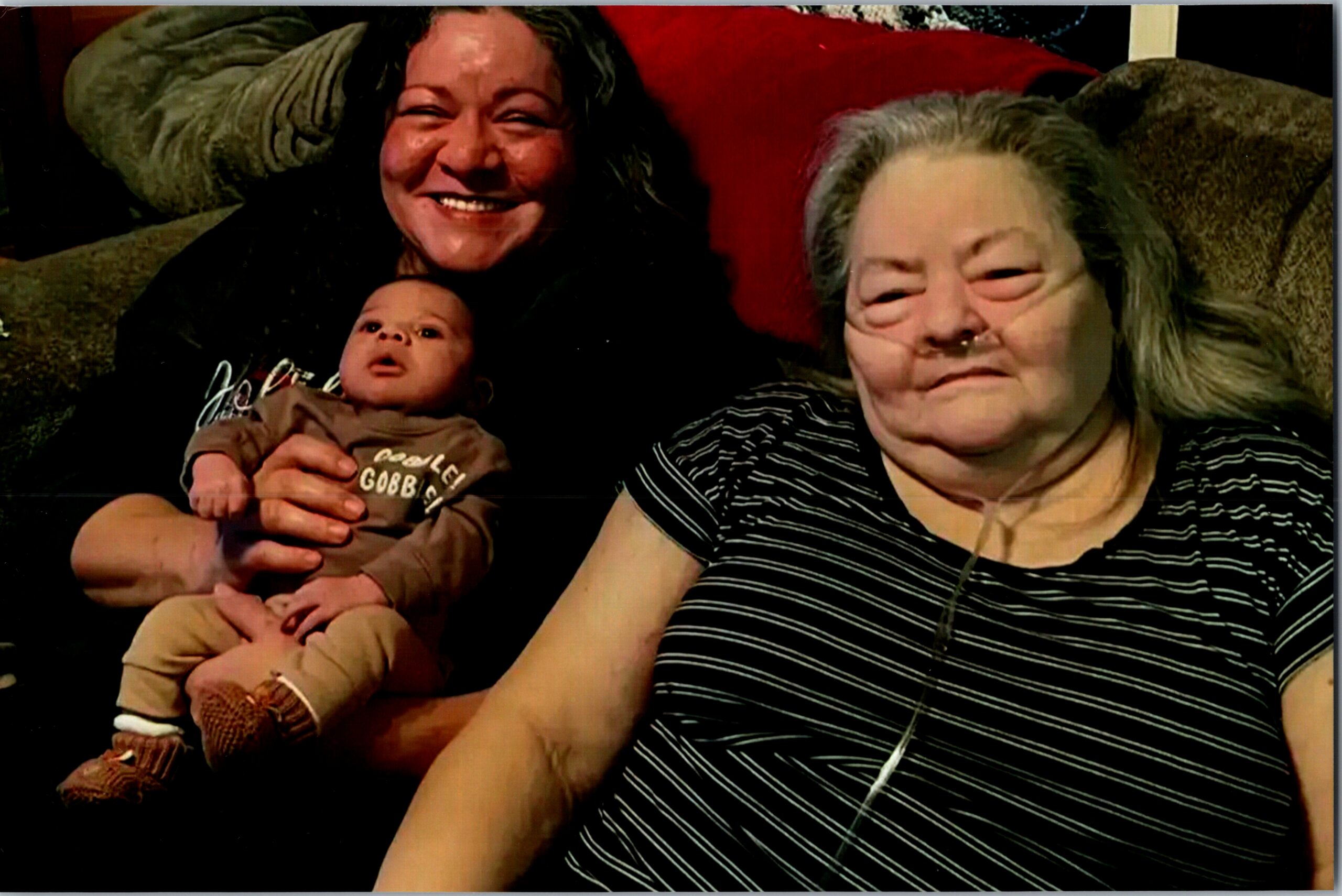 Smiling woman holding a baby on her lap, seated with an older woman on a couch.