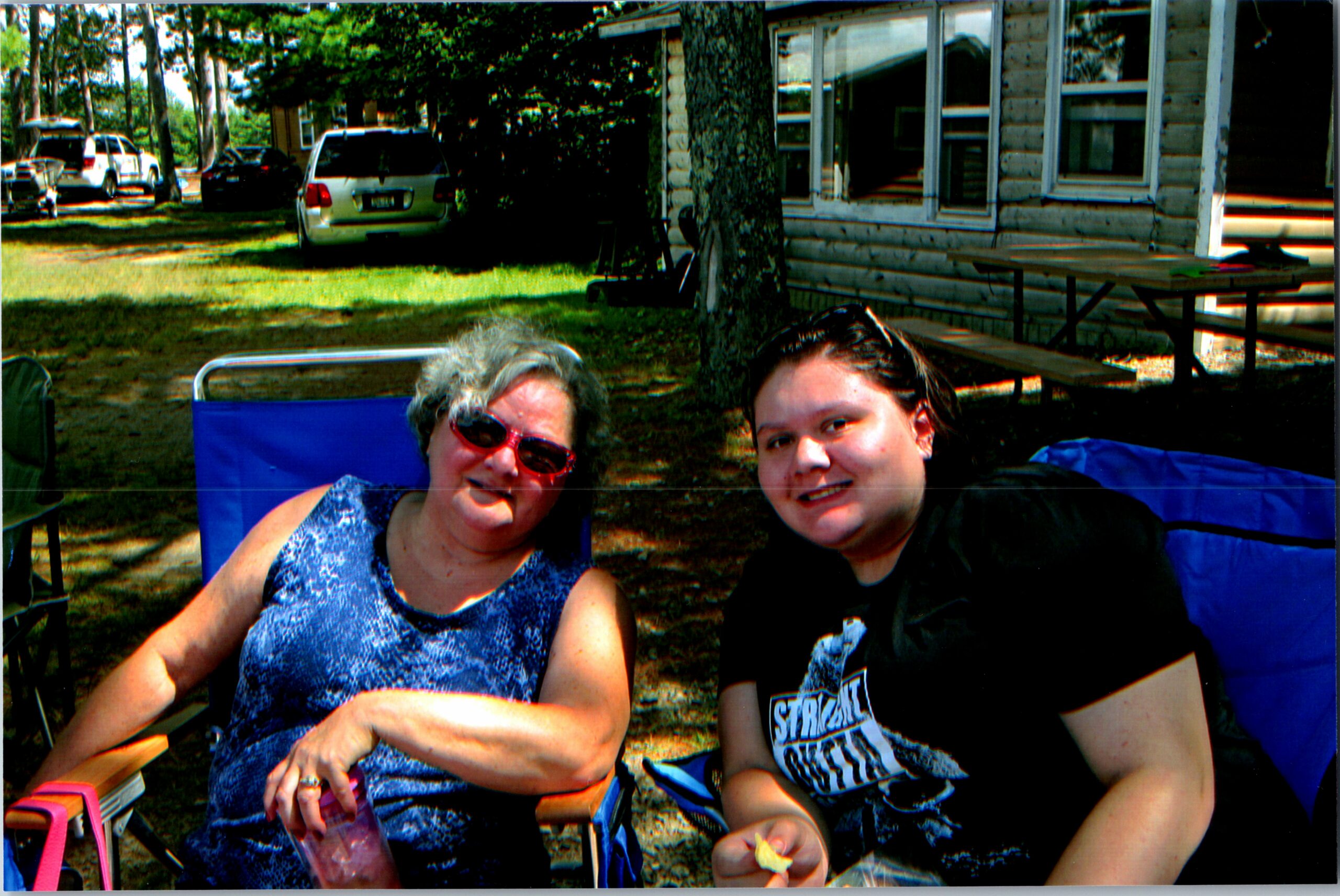 Two women smiling while sitting in blue camping chairs outdoors, with a wooden house, trees, and parked cars in the background.