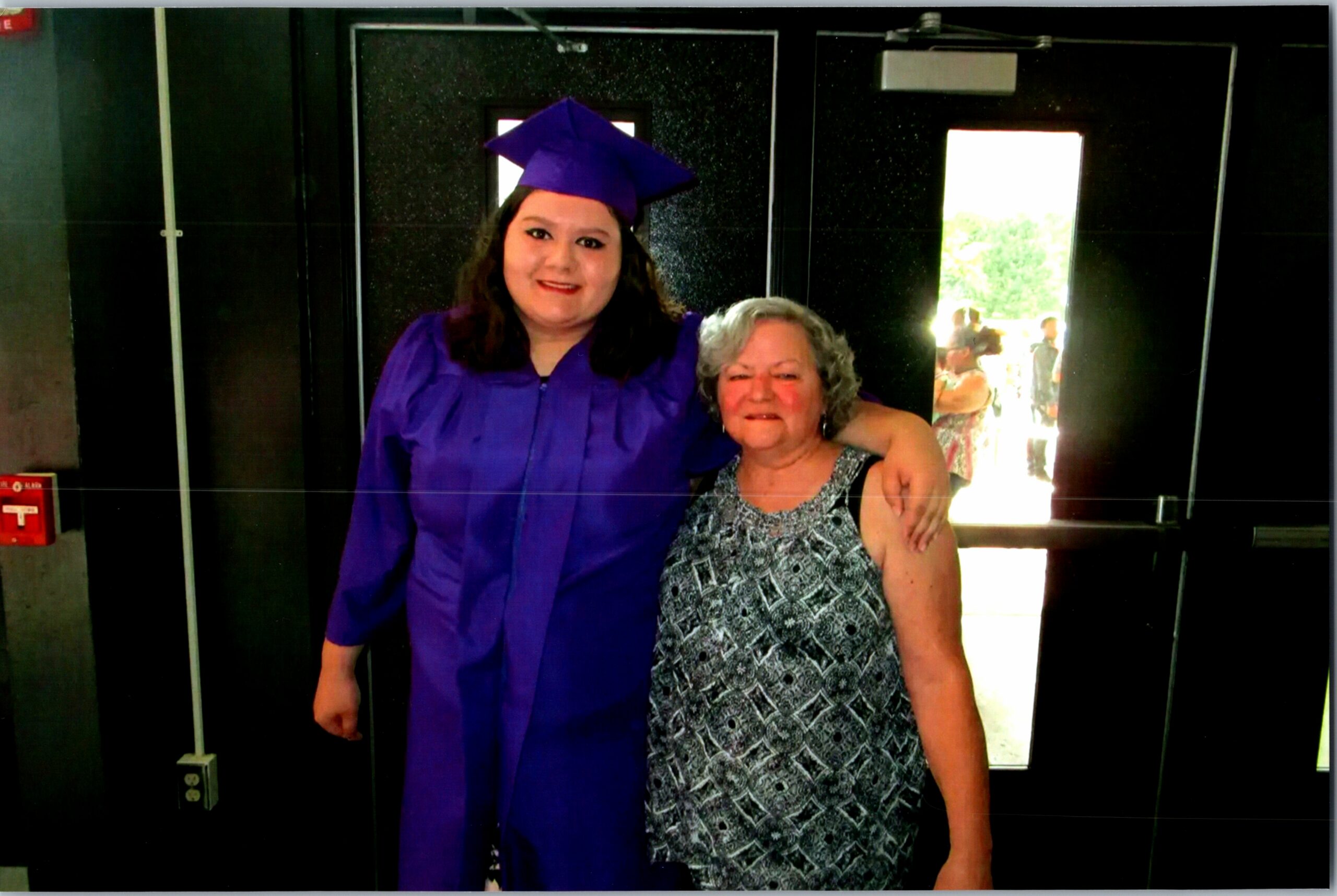 Young graduate in purple cap and gown posing with an older woman in a patterned dress inside a building, smiling at the camera.