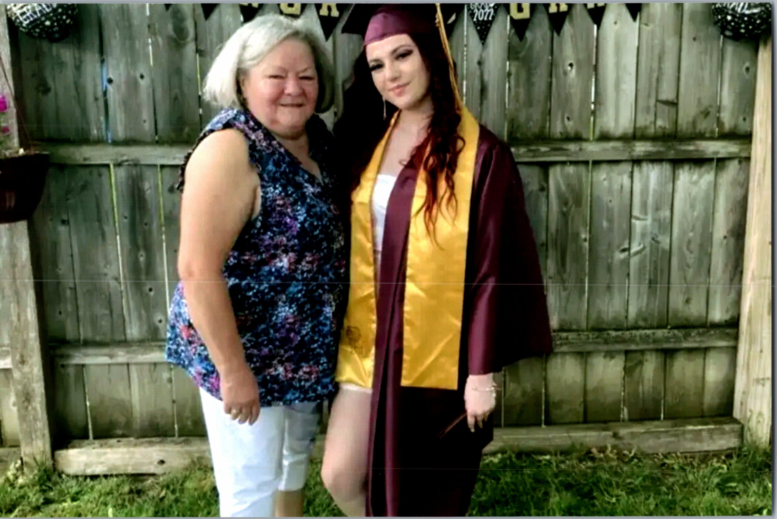 Graduate in burgundy cap and gown with a yellow stole posing with a smiling older woman in a floral top, against a wooden fence backdrop.