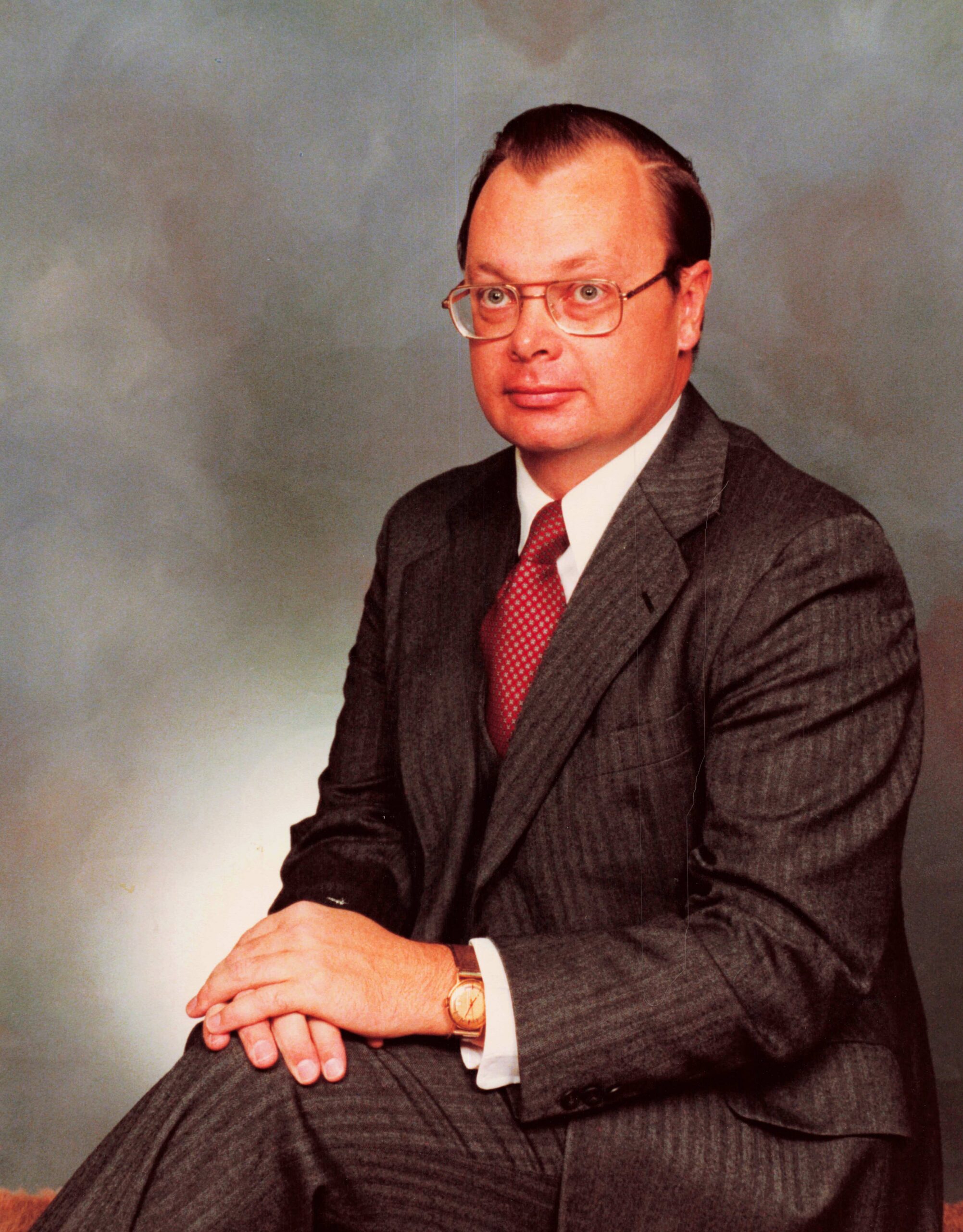 Formal portrait of a middle-aged man in a pinstripe suit and red tie, wearing glasses, with hands folded in his lap in a studio setting.