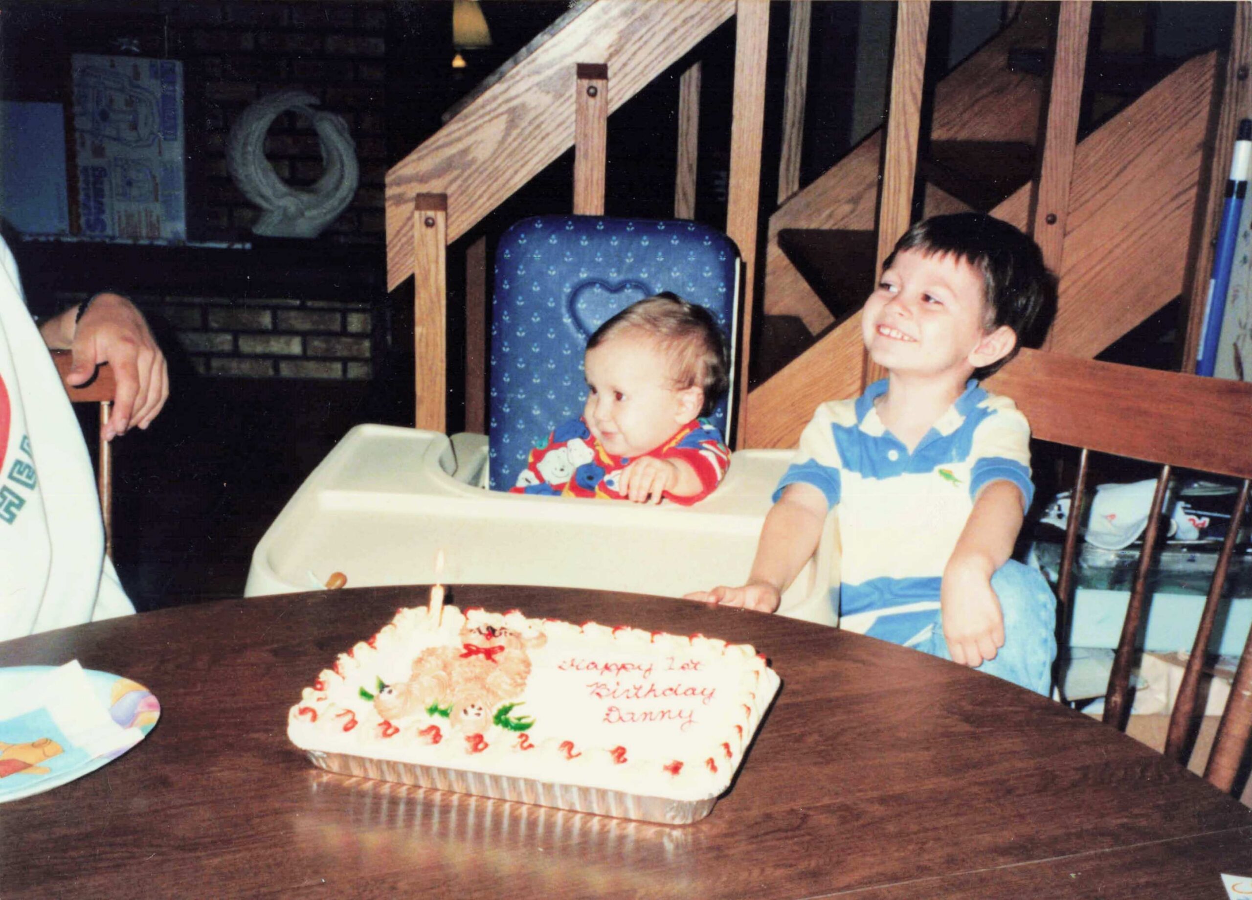 Two young children sit in high chairs at a birthday party, cake with candles in front; older boy smiles nearby.