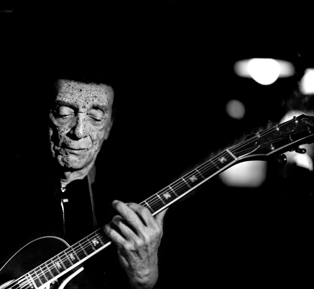 Black-and-white photo of an elderly man playing an acoustic guitar with his eyes closed, focused on the music.