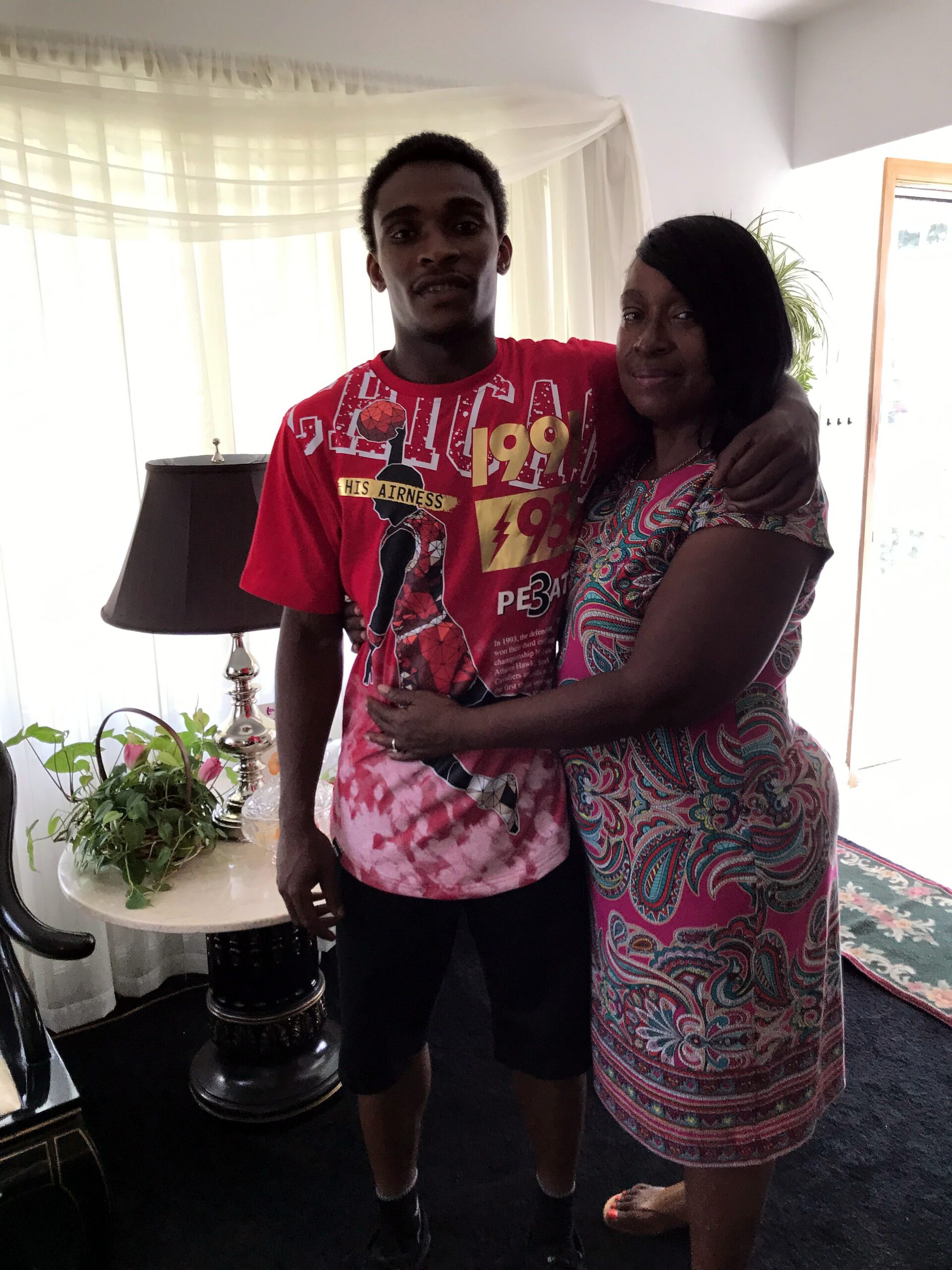 Two people posing together indoors with arms around each other; man in a red graphic T-shirt and woman in a colorful patterned dress, near a lamp and plants in a living room.