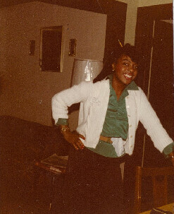 Woman in a green blouse and white jacket stands with hands on hips, smiling in a living room.