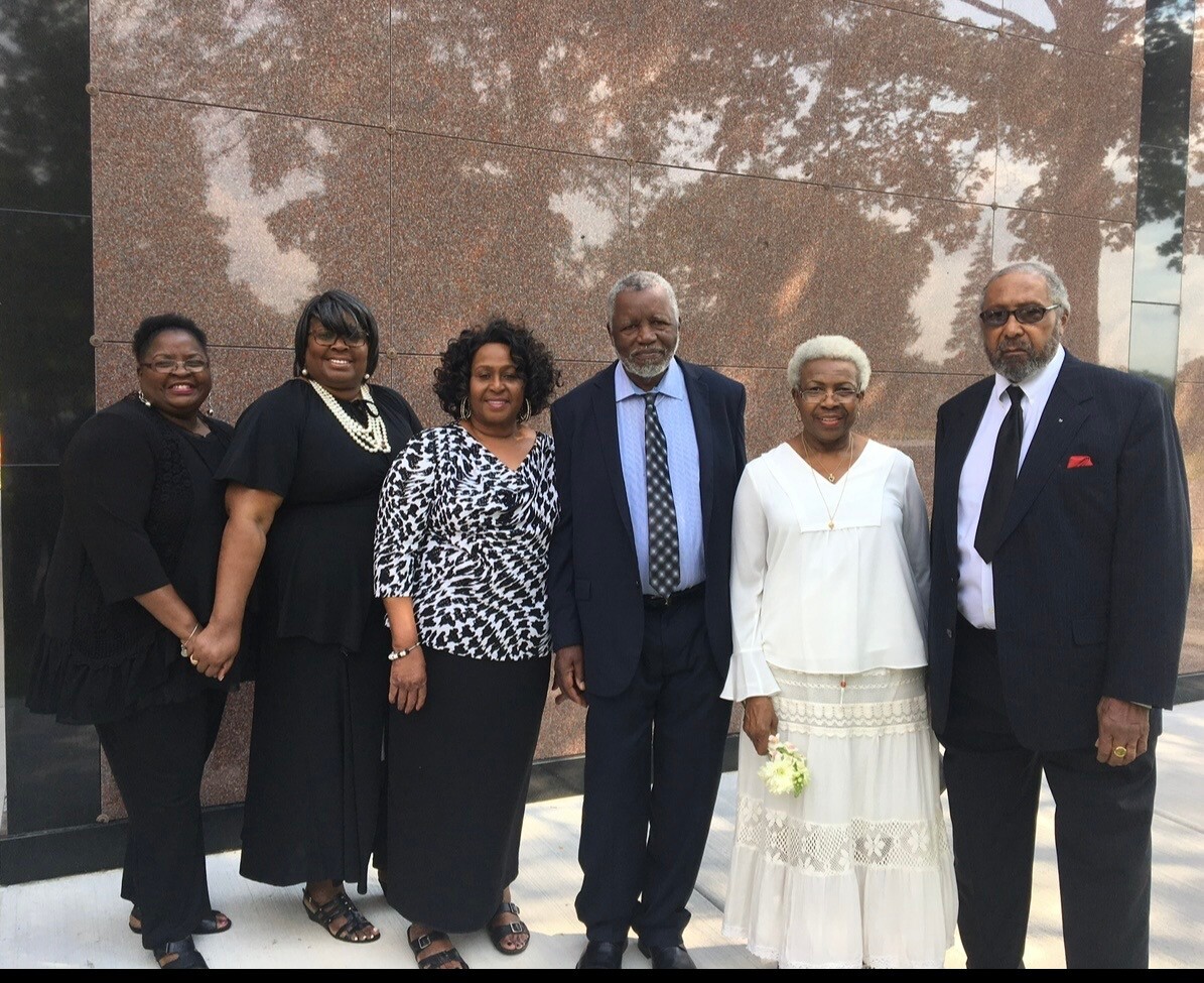 Group of six adults in formal attire posing together outdoors against a brown granite wall.