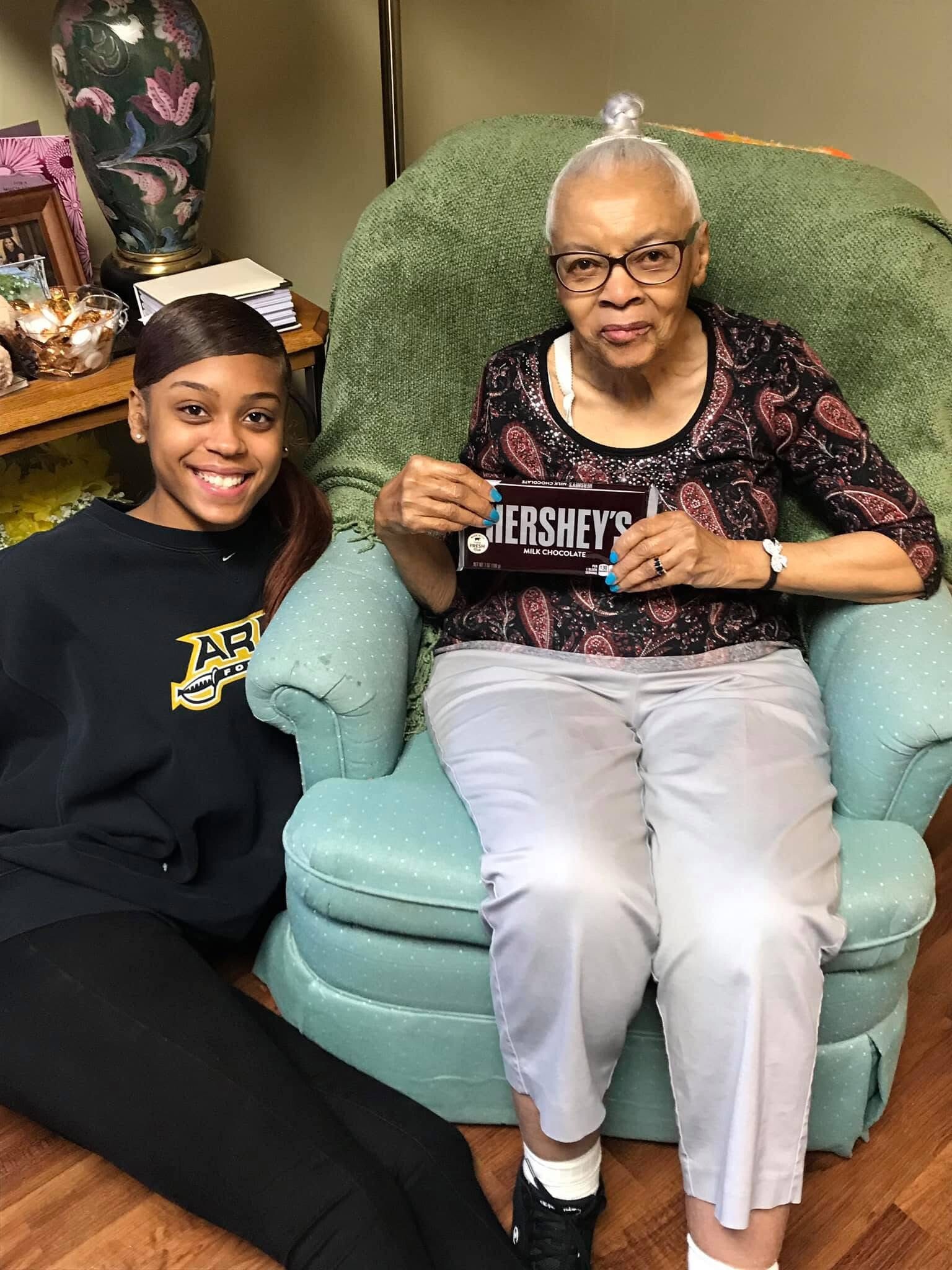 Smiling young woman sits on the floor beside an elderly woman in a teal upholstered chair, who holds a Hershey's milk chocolate bar.