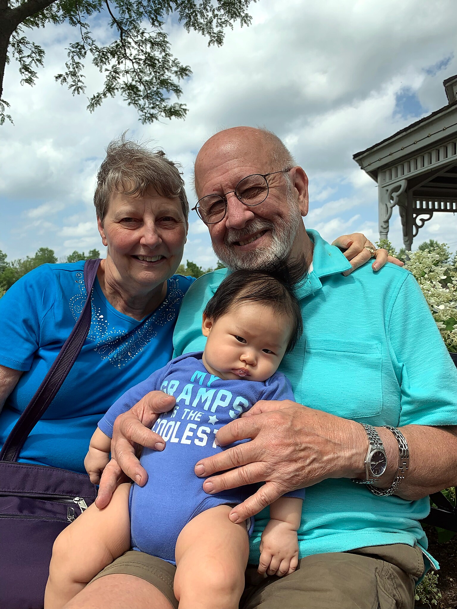 Smiling older couple holding a baby outdoors on a sunny day.