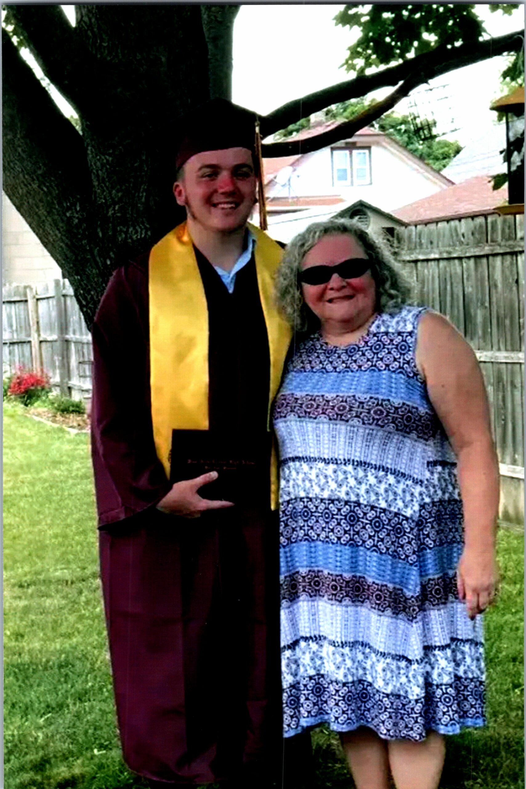 Young graduate in a maroon gown and yellow stole posing with a smiling woman in a backyard.
