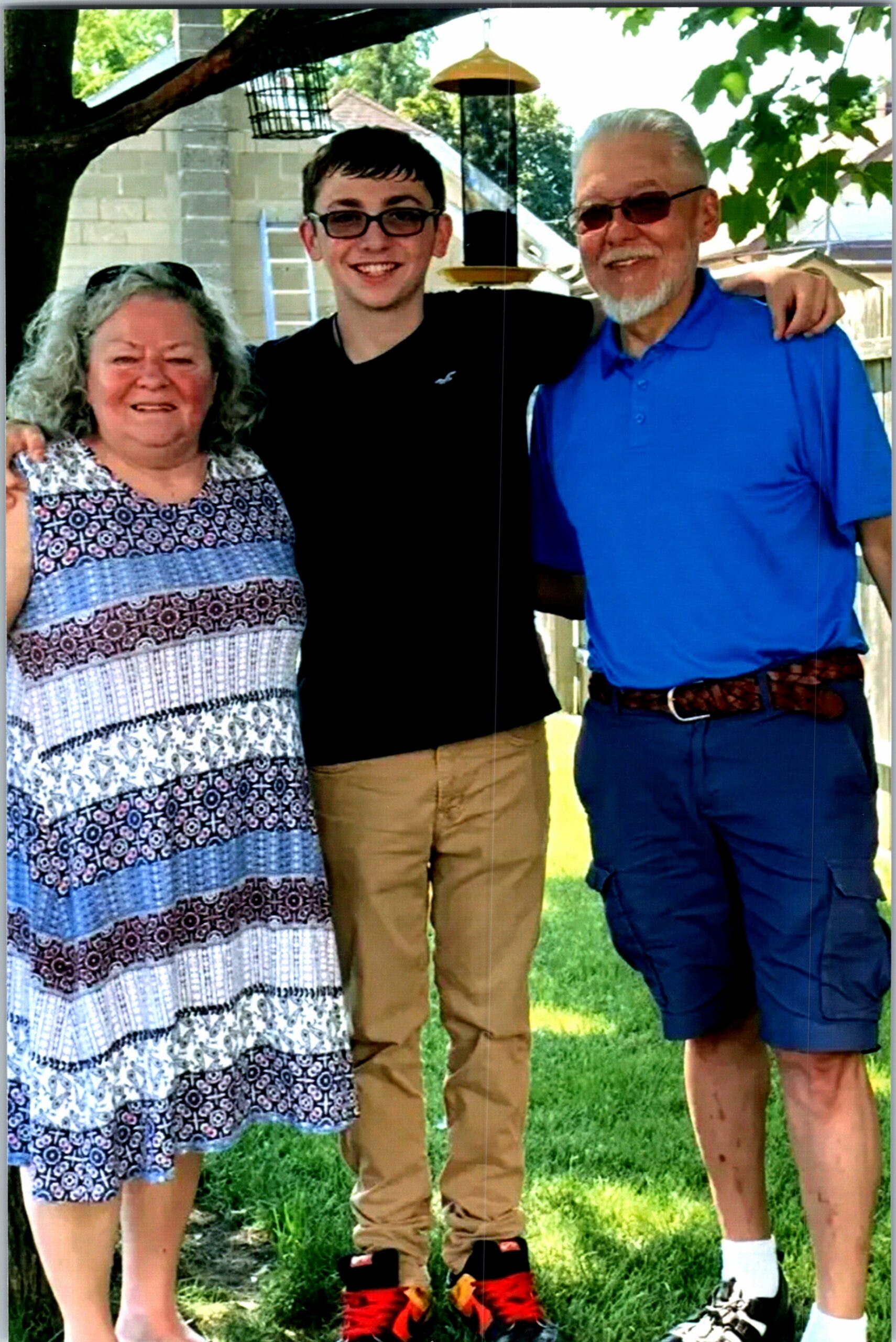 Three generations posing together outdoors, smiling with arms around each other under a tree in a sunny yard.