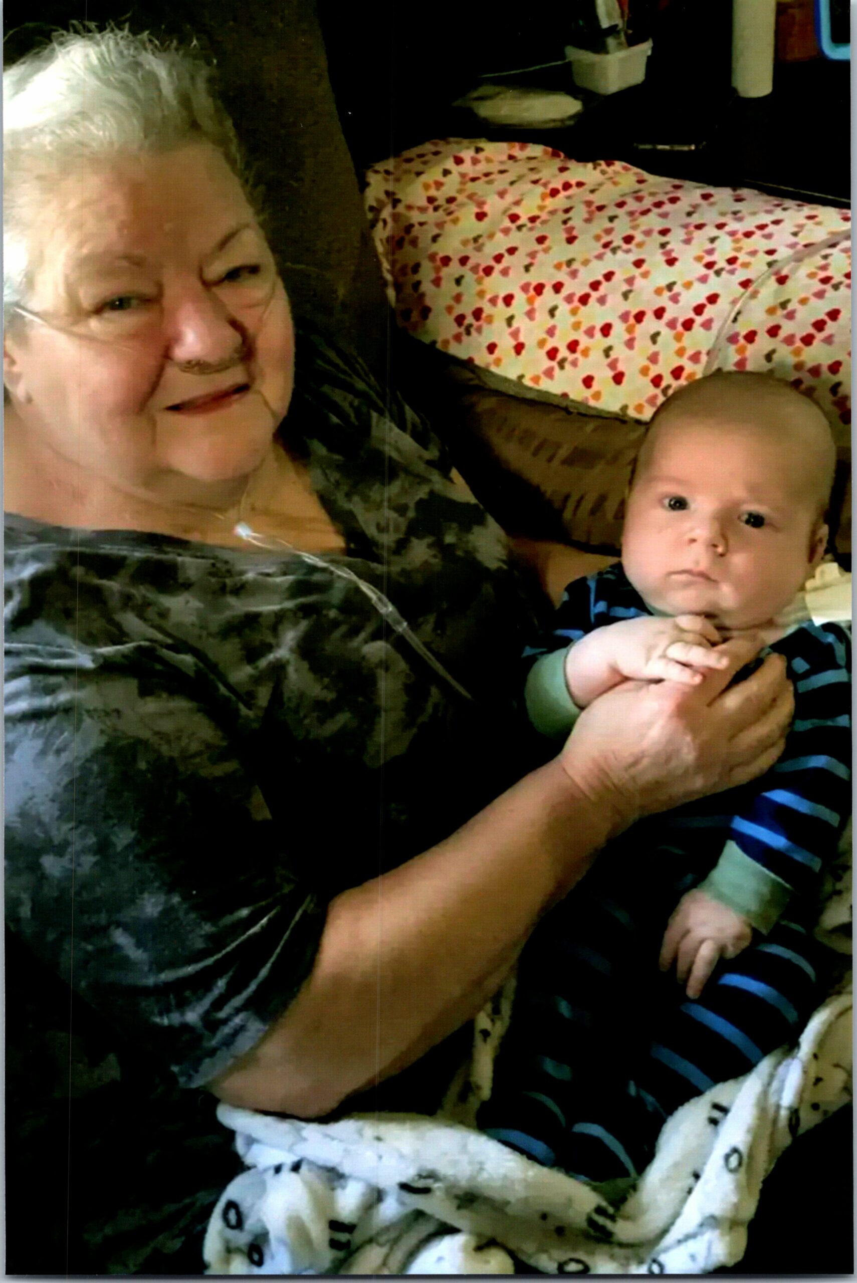 Elderly woman with an oxygen tube smiling while holding a baby in her arms at home.