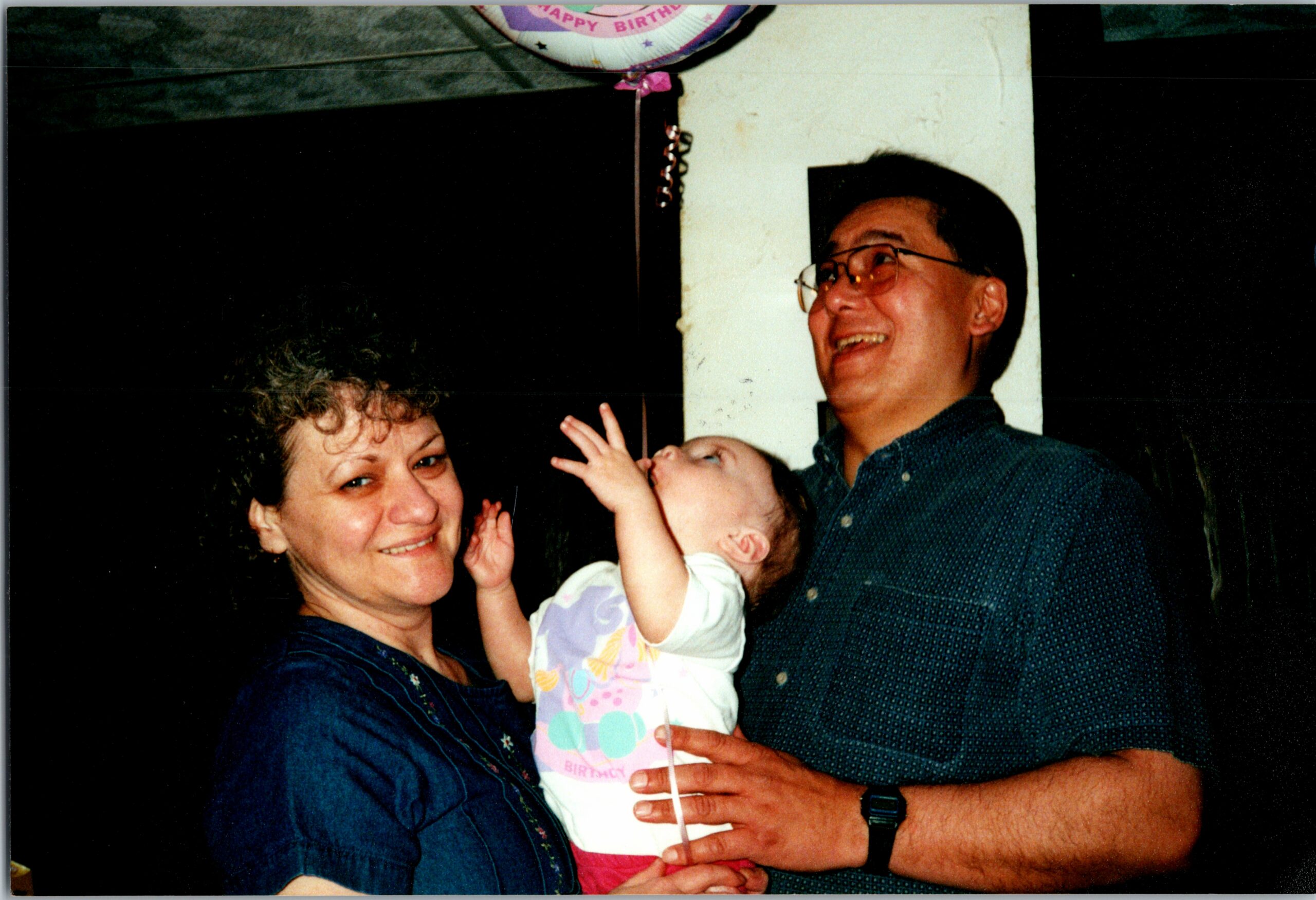 Mother and father smiling while holding a baby during a birthday celebration, with a balloon visible above them.