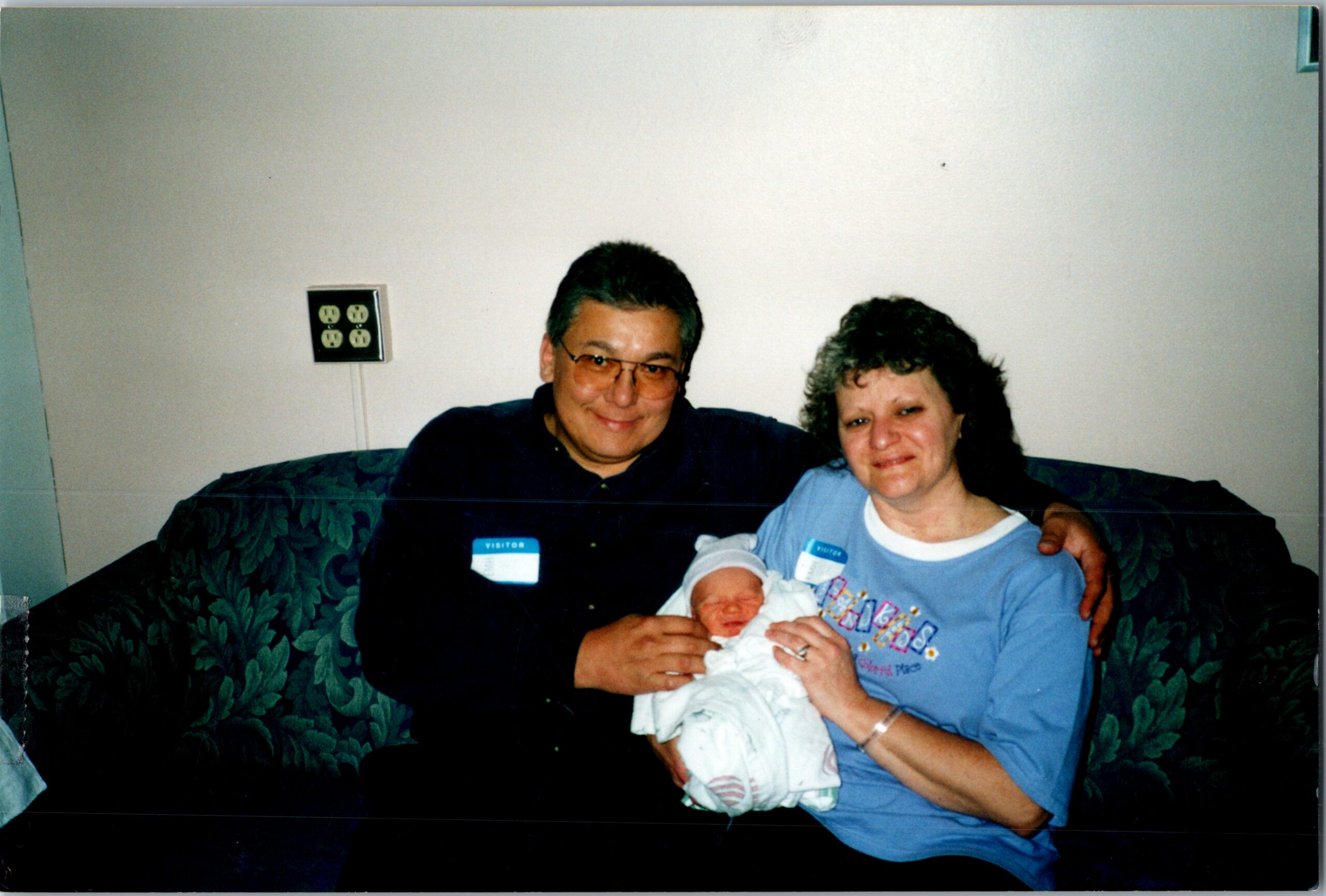 Smiling couple sitting on a patterned couch, cradling their newborn baby wrapped in a white blanket in a hospital/visit setting.