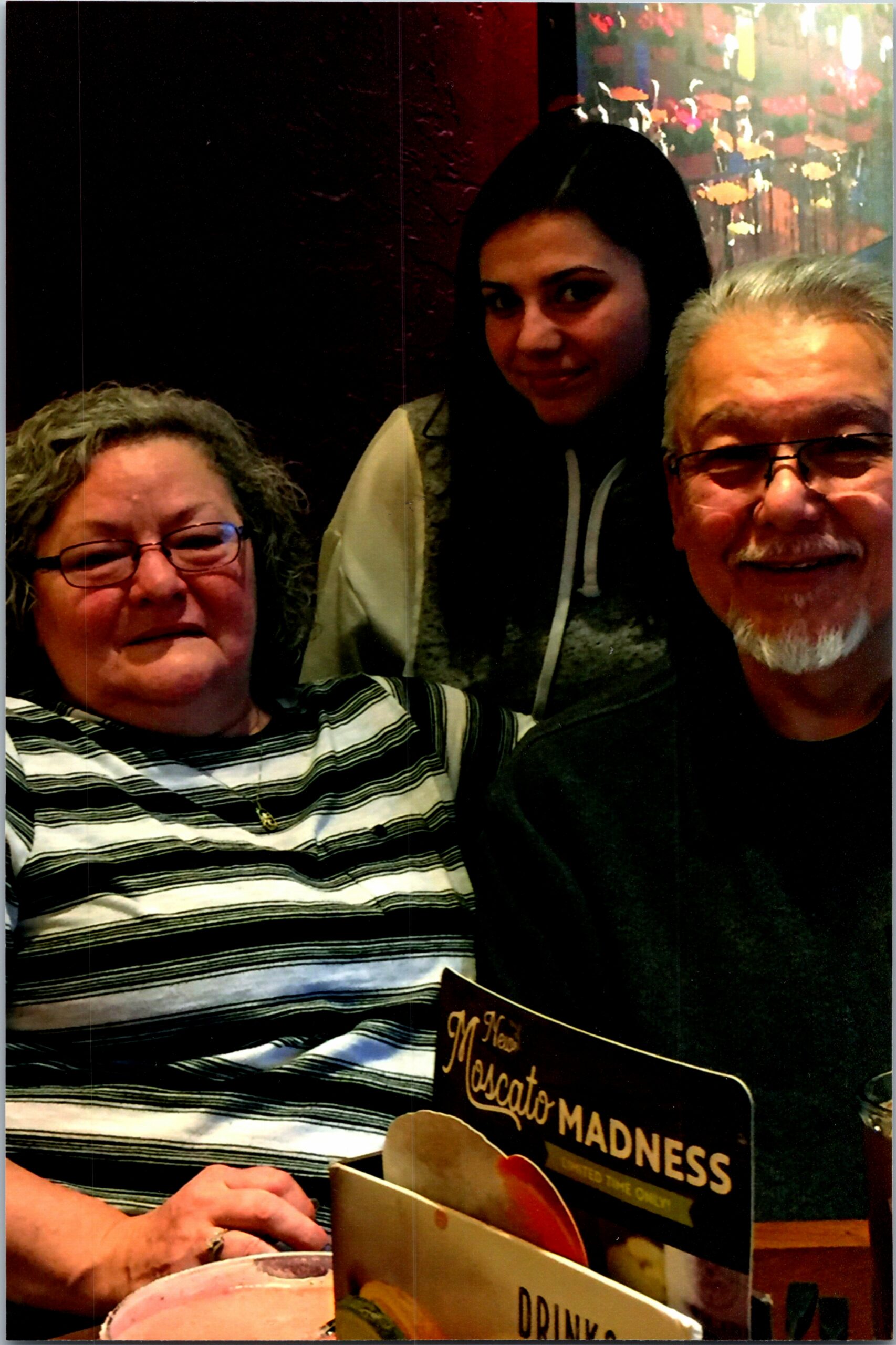 Three people sit close together smiling at the camera in a restaurant, with a menu visible in the foreground.
