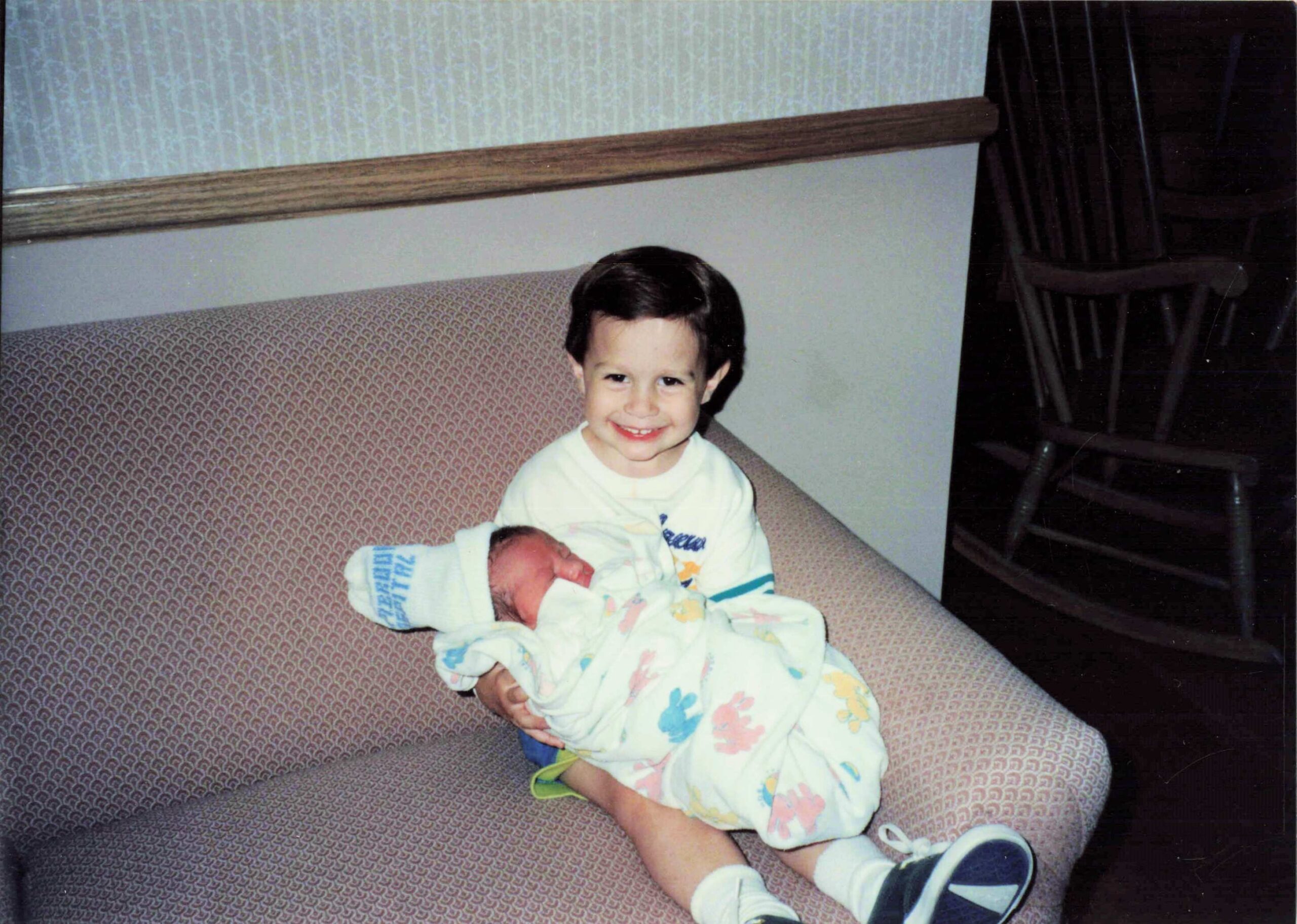 Two young siblings on a patterned sofa, the older one smiling while cradling a newborn baby wrapped in a pastel blanket te baby close to the chest