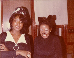 Two women pose for a casual photo indoors; one wears a hat with a star and a large ring, the other has dark hair styled with bear-ear clips.