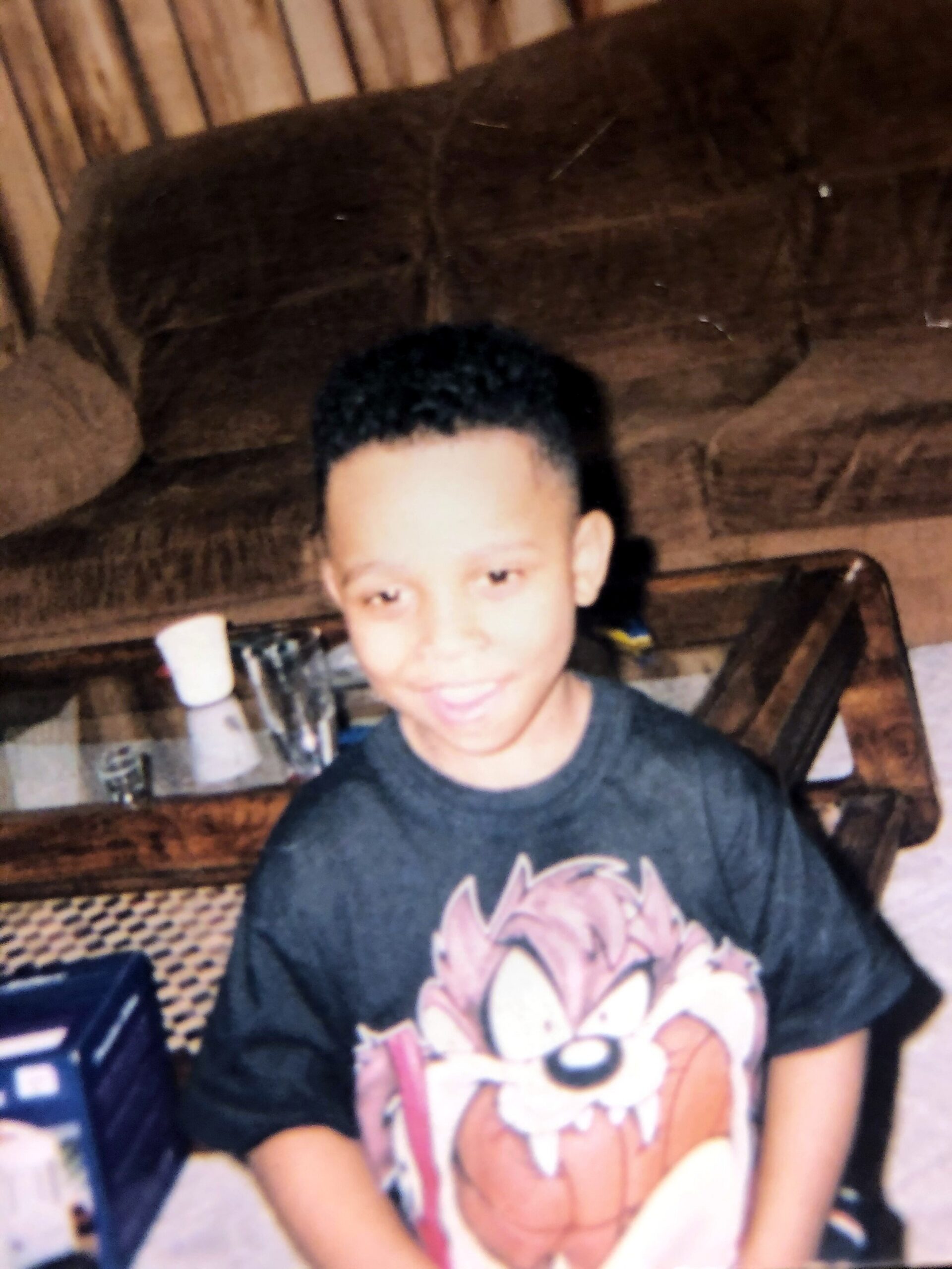 Smiling boy with short black hair wearing a dark graphic tee, seated on a brown couch in a wood-paneled room.